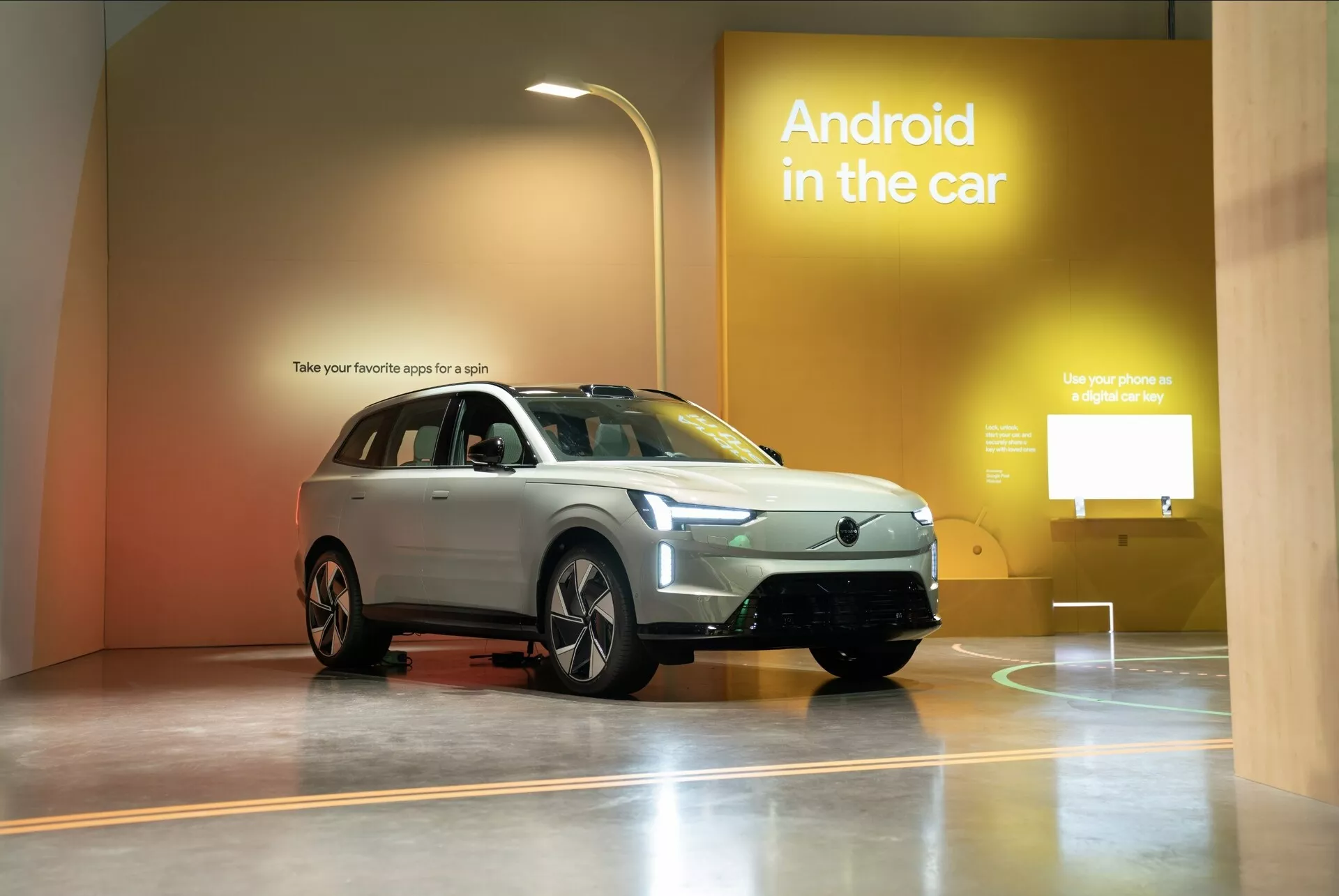 Volvo EX90 prve generacije 2023 razstavljeno v razstavnem salonu, ki poudarja njegov SUV slog, pogled spredaj tri četrtine vozila, ki prikazuje rešetko, žaromete, sprednji odbijač, kapuco, kolesa, stranska ogledala in okna z reklamo za tehnologijo Android v avtu v ozadju
