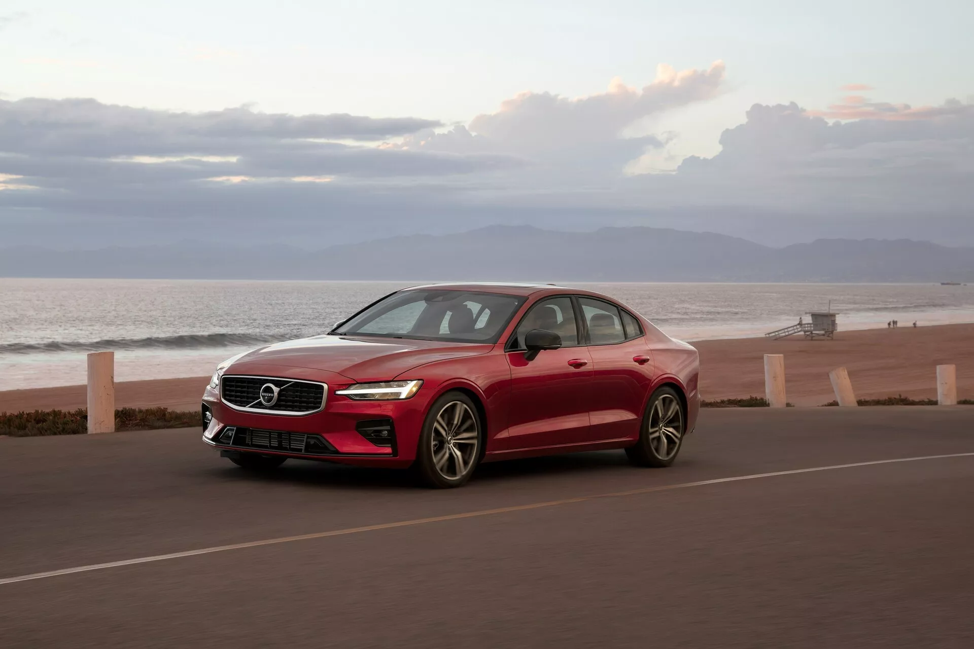 Volvo S60 limuzina 2018 s tričetrtinskega sprednjega kota na obalni cesti, živahno rdeča, z oceanom v ozadju. Značilnosti vključujejo eleganten aerodinamičen dizajn, LED dnevne luči, večkraka lita platišča, karoserijska ogledala in strešno okno. Posneto v jutranji ali večerni svetlobi.