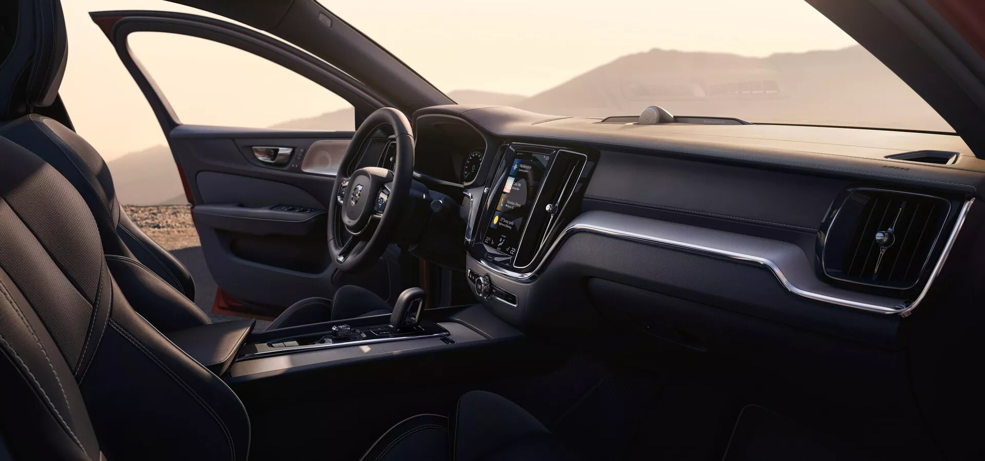 Notranjost avtomobila Volvo S60/V60 2018 (3. generacija) s perspektive sopotnika. Slika prikazuje sprednjo kabino, vključno z armaturno ploščo, volanom, sredinsko konzolo in delom sopotnikovega sedeža. Armaturna plošča ima velik centralno nameščen zaslon na dotik za informacijsko-razvedrilni sistem in minimalne fizične kontrole, kar poudarja eleganten, moderen dizajn. Volan je usnjen z vgrajenimi kontrolami, sedeži pa so oblazinjeni z vidnimi šivi. Ozadje, vidno skozi okna, nakazuje zunanjo lokacijo s hribi ali gorami.