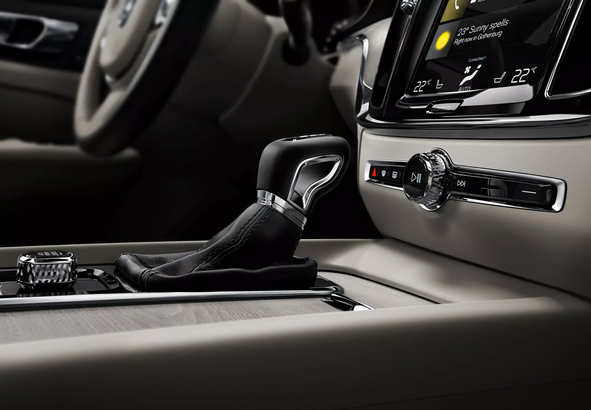 Podroben posnetek notranjosti vozila Volvo S60-V60 2018 (3. generacija), osredotočen na področje osrednje konzole. Eleganten prestavni ročaj je v navpičnem položaju, v parkirnem ali nevtralnem položaju, oblečen v usnje z kovinskimi dodatki. V ozadju je prikazan zaslon na dotik infotainmenta, ki prikazuje dvopodročno klimatsko napravo nastavljeno na 22°C, rahlo nagnjen proti vozniku. Desno od prestavnega ročaja je gumb za zagon/zaustavitev, ki omogoča zagon brez ključa. Na levi strani je nadzorna ročica in gumbi za navigacijo po menijih infotainmenta. Konzola je okrašena s kakovostnim lesom in brušenim kovom, kar odraža luksuz in mojstrsko izdelavo vozila.
