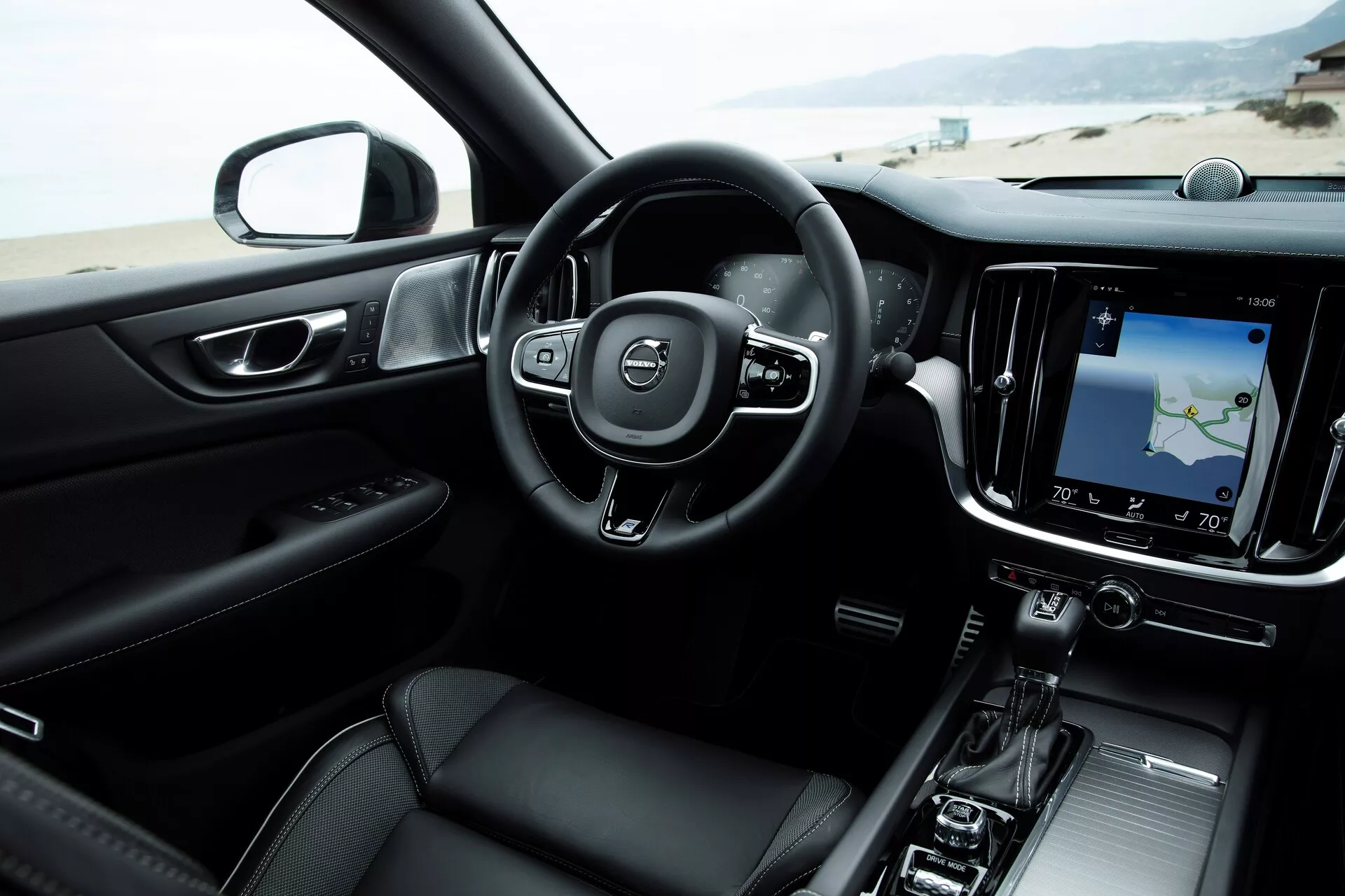 Notranji pogled na sodoben Volvo S60-V60 2018 (3. generacija) iz perspektive sprednjega sovoznikovega sedeža, s trikraka volanom z vgrajenimi kontrolami, digitalnimi instrumenti, velikim pokončnim zaslonom na dotik infotainmentnega sistema z navigacijskim zemljevidom, minimalističnimi nadzornimi enotami HVAC, elegantno prestavno ročico, prestižno temno barvno shemo z vstavki iz kovine in naprednim avdio sistemom.
