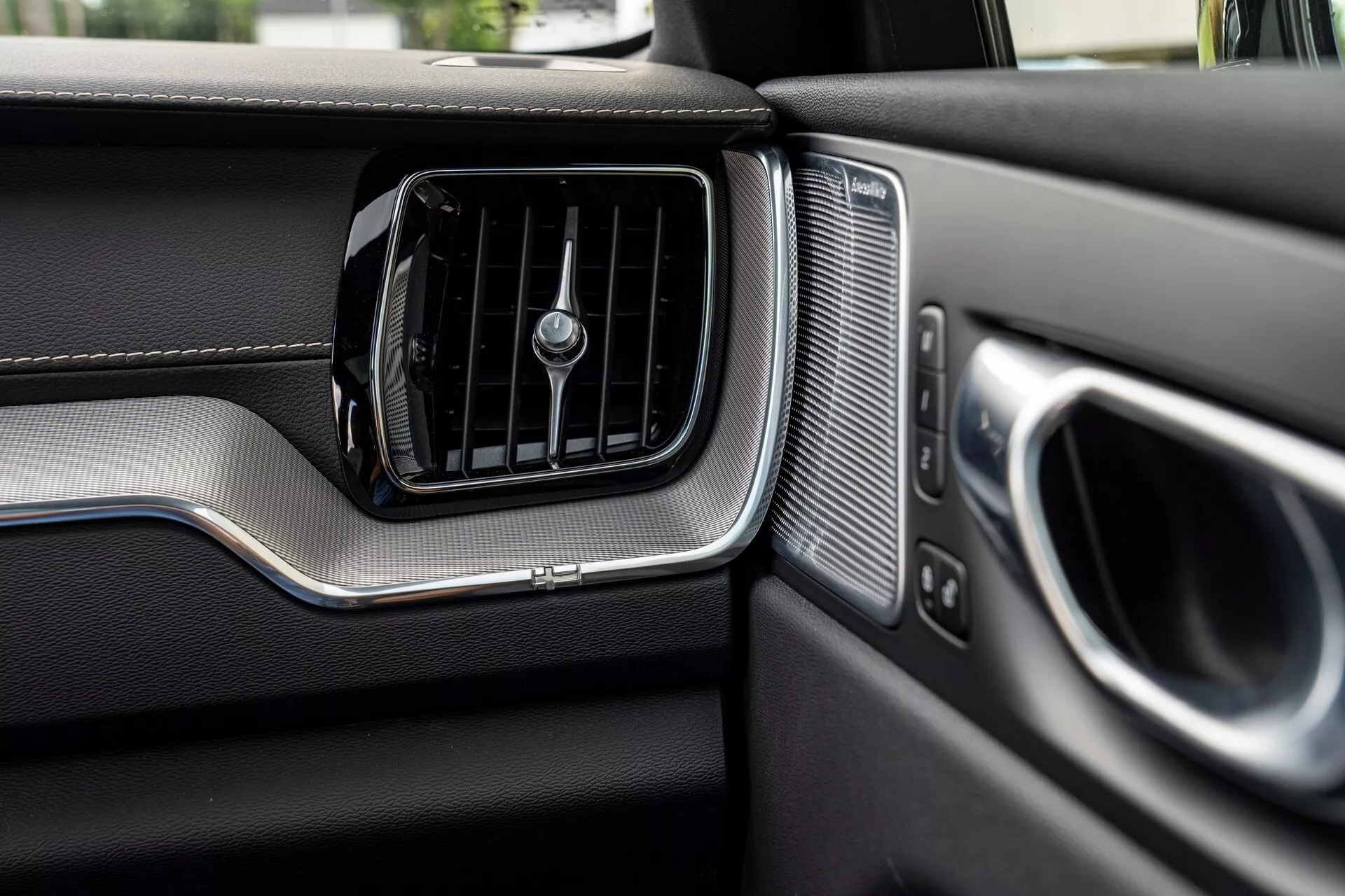 Podroben pogled na armaturno ploščo na strani sopotnika v vozilu Volvo XC60 2021 (2. generacija facelift). Slika jasno prikazuje zračno odprtino za klimatsko napravo s horizontalnimi režami in sredinskim vrtljivim gumbom, obrobljenim s kovinskim poudarkom. Desno od zračne odprtine se nahaja rešetka zvočnika z enakomernimi perforacijami, poleg delno vidnega naslona za roko na vratih in ročaja, prav tako z kovinskimi poudarki. Armaturna plošča prikazuje materiale, podobne usnju, na zgornjem delu in drugače strukturiran spodnji del, ki je ločen s kovinskim trakom, kar poudarja vrhunski dizajn in izdelavo vozila.