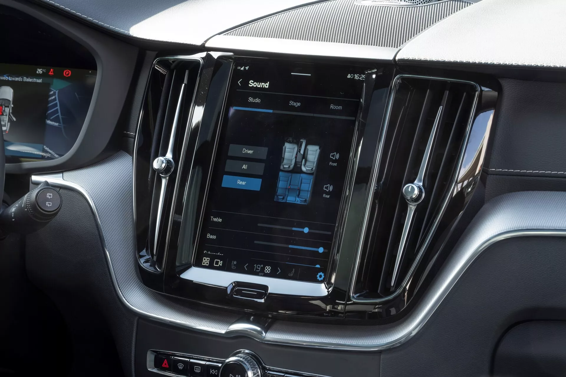 Notranji posnetek Volva XC60 2021 facelift, osredotočen na sredinsko konzolo z navpično postavljenim velikim zaslonom na dotik infotainment sistema. Posnet iz perspektive potniškega sedeža, slika prikazuje tudi nastavitve dvopodročnega klimatskega nadzora, kovinske prezračevalne reže in ročne upravljalne gumbe pod zaslonom, kar daje vtis sodobnega in luksuznega dizajna.