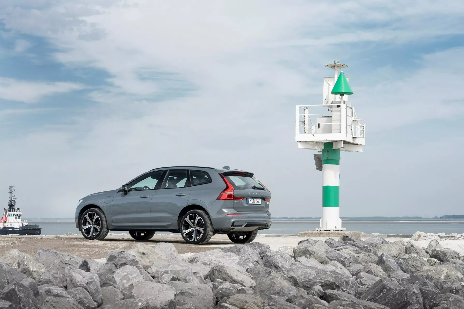 Fotografija modela Volvo XC60 2021 druge generacije s prenovo, posneta iz tristranskega zadnjega kota na skalnati površini, z morskim ozadjem. Temno sivo avtomobil s kovinskim zaključkom ima elegantno, športno strešno linijo, večkraka aluminijasta platišča, zadnje parkirne senzorje in dvojni izpuh. Na desni strani avtomobila je viden majhen svetilnik ali signalna luč, z vlačilcem ali pilotskim čolnom v vodi, kar poudarja obalno vzdušje.