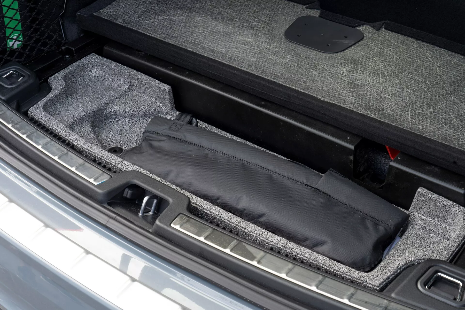 Podroben pogled na prtljažni prostor Volvo XC60 2021, ki prikazuje skrit predal pod dnom prtljažnika s črnim ovojem, notranjost s preprogo in vidnim kovinskim pragom, poleg rdečega orodja za nujne primere.