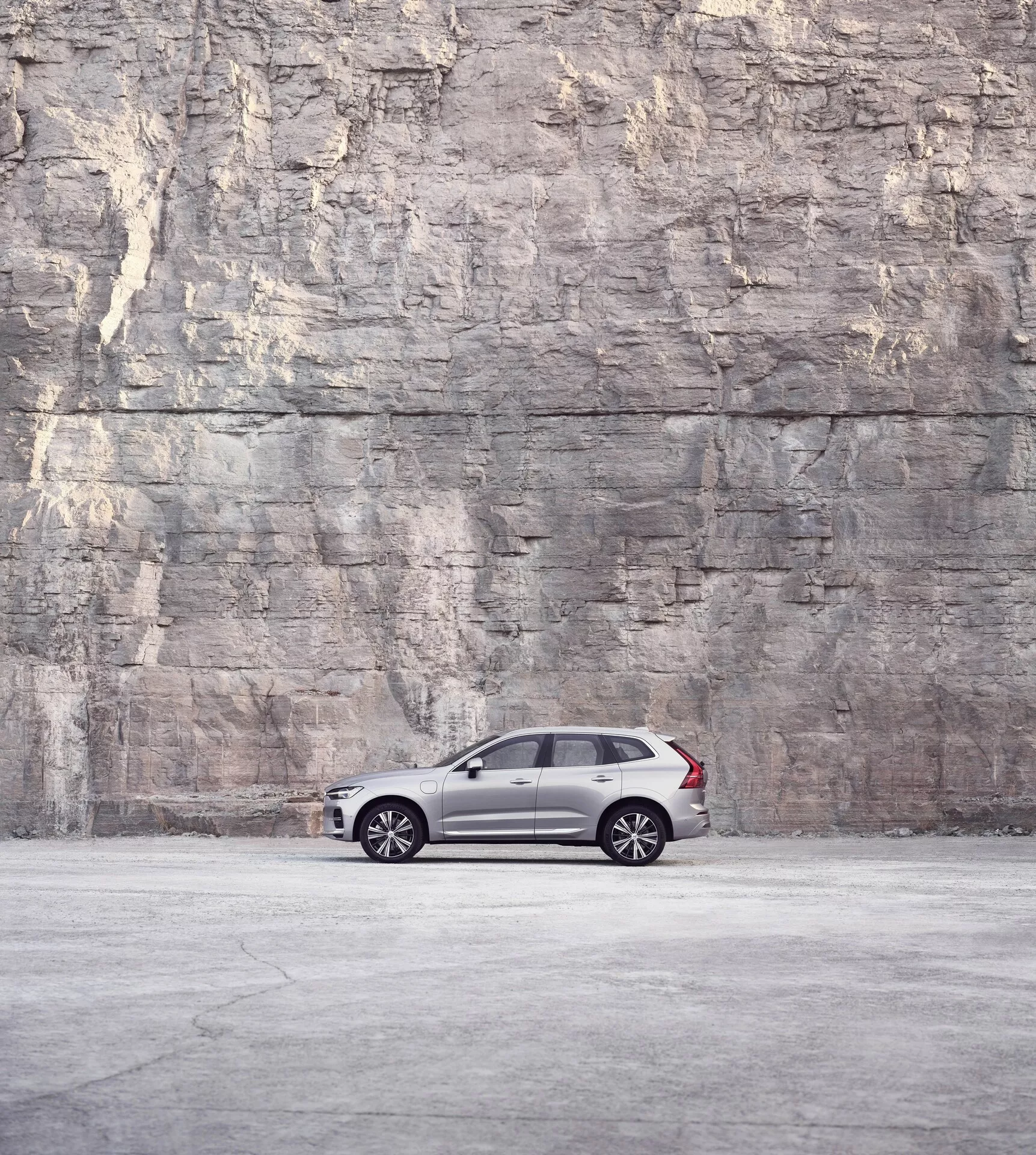 Poglej s strani na Volvo XC60 2021 (2. generacija facelift), parkiranega pred veliko teksturirano steno iz kamna ali betona, ki prikazuje svojo elegantno in luksuzno zasnovo s temno kovinsko barvo, platišči z več kraki, zatemnjenimi okni in črnimi B-stebrički pod mehko, difuzno svetlobo.