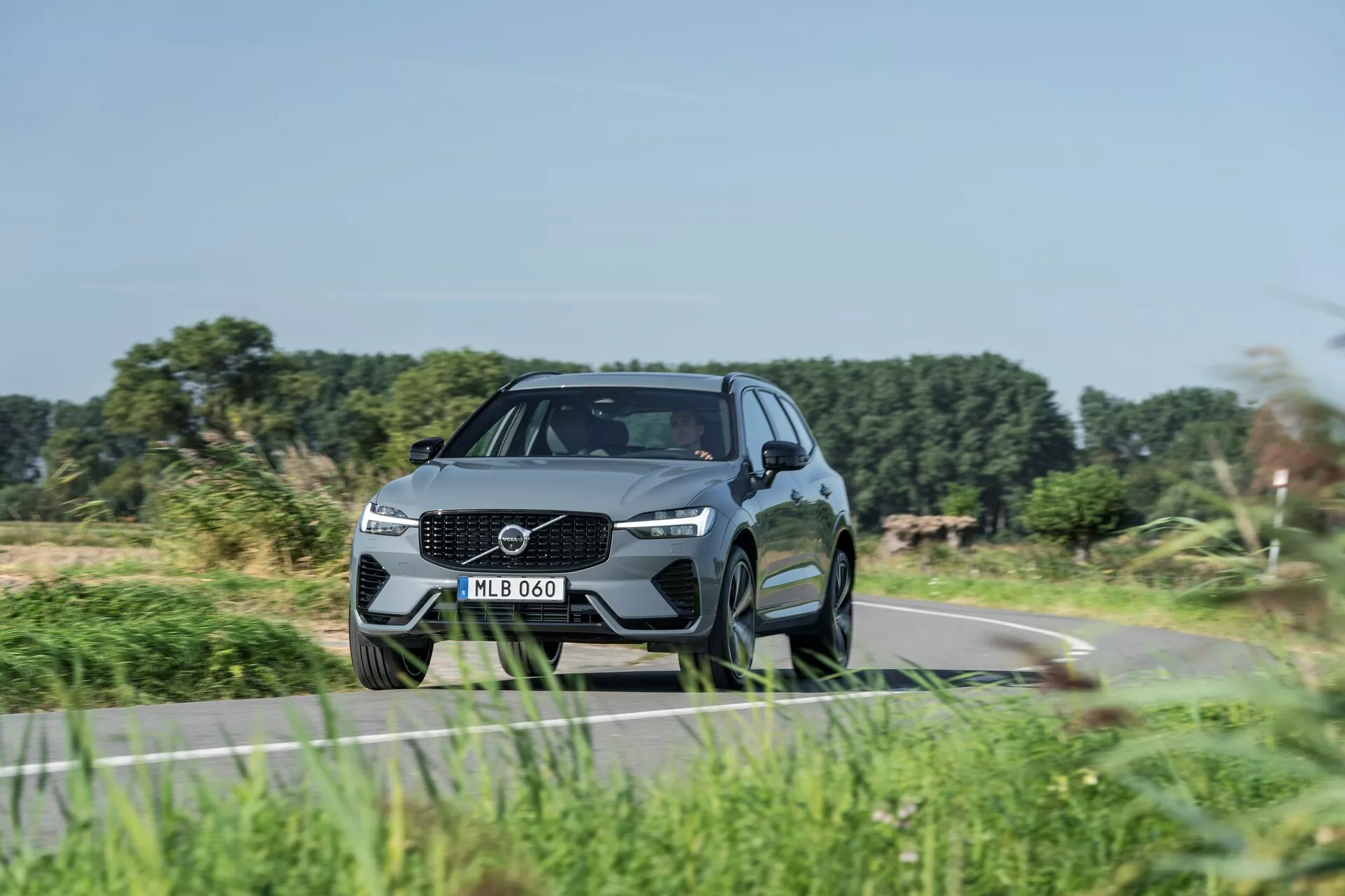 Modern SUV, Volvo XC60 2021 (2. generacija facelift), ujet iz nizkega kot prednje tretjine, ki poudarja dinamični položaj in oblikovne značilnosti. Značilna zasnova rešetke z velikim emblemom, elegantne LED žaromete in oblikovan odbijač z integriranimi zračnimi odprtinami. Aerodinamičen, visokokakovosten videz s profiliranimi linijami, visoko oddaljenostjo od tal, velikimi večkrakimi aluminijastimi platišči, nizkoprofilnimi pnevmatikami, stranskimi ogledali z vgrajenimi smerniki, poševno strešno linijo in zatemnjenimi stekli. Nakazuje sodobne tehnološke in varnostne funkcije, vozi se po podeželski cesti z zamegljeno vegetacijo, kar nakazuje gibanje.