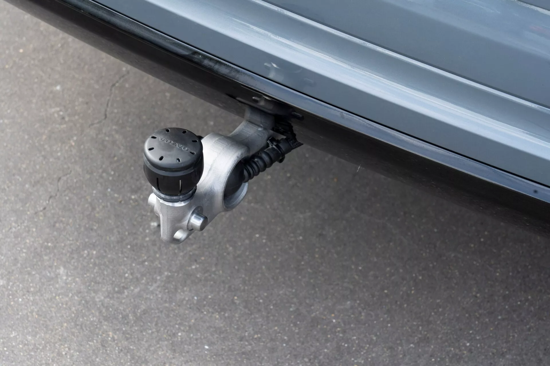 Približan posnetek izvlečnega stranskega stopalca na vozilu Volvo XC60 2021 (2. generacija-facelift), ki prikazuje razširjeno nedrsečo površino z izbočenimi črnimi pikami pod stranskimi vrati vozila, pritrjeno na mehanizem za tečaje iz kovine. Fotografirano iz nizkega kota, poudarja avtomatizirano funkcijo, zasnovano za lažje vstopanje in izstopanje z zmanjšanjem razdalje do tal.
