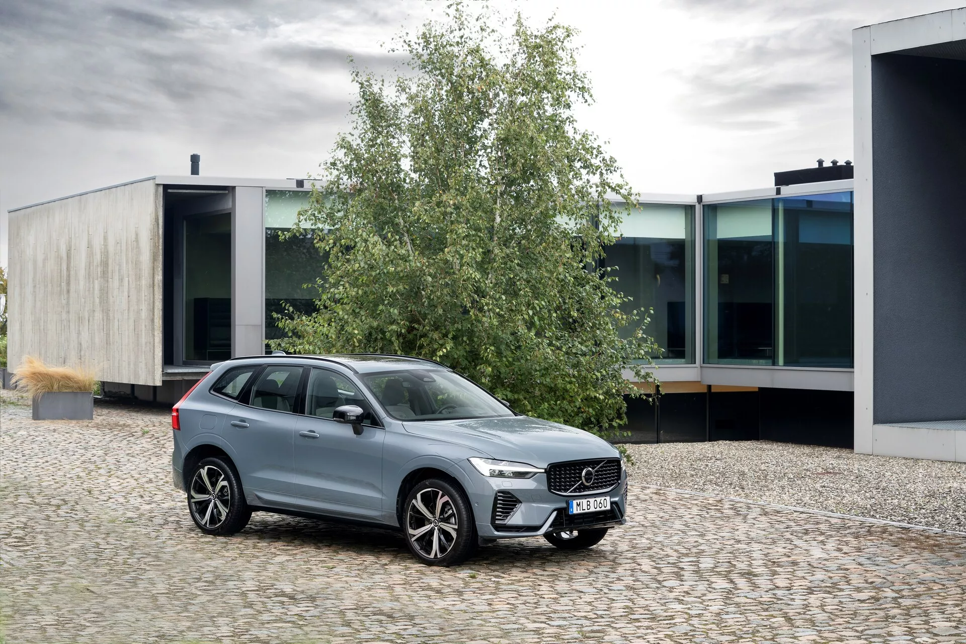 Slika Volva XC60 2021 (2. generacija - facelifting) parkiranega na tlakovani površini, postavljenega pod rahlim kotom proti levi. Sprednji tričetrtinski pogled poudarja izrazito rešetko z navpičnimi palicami, elegantnimi LED žarometi z dnevnimi lučmi in velikimi platišči iz lahke zlitine z večkrakim oblikovanjem. Okna so obdana s kromom, kar povečuje njegov poliran kovinski videz. SUV ima povečano višino vožnje, strešne letve in barvno usklajene stranske ogledala z vgrajenimi smerniki. Scena se odvija v urbanem okolju z moderno arhitekturo, ki prikazuje eleganten dizajn in luksuzne lastnosti vozila.