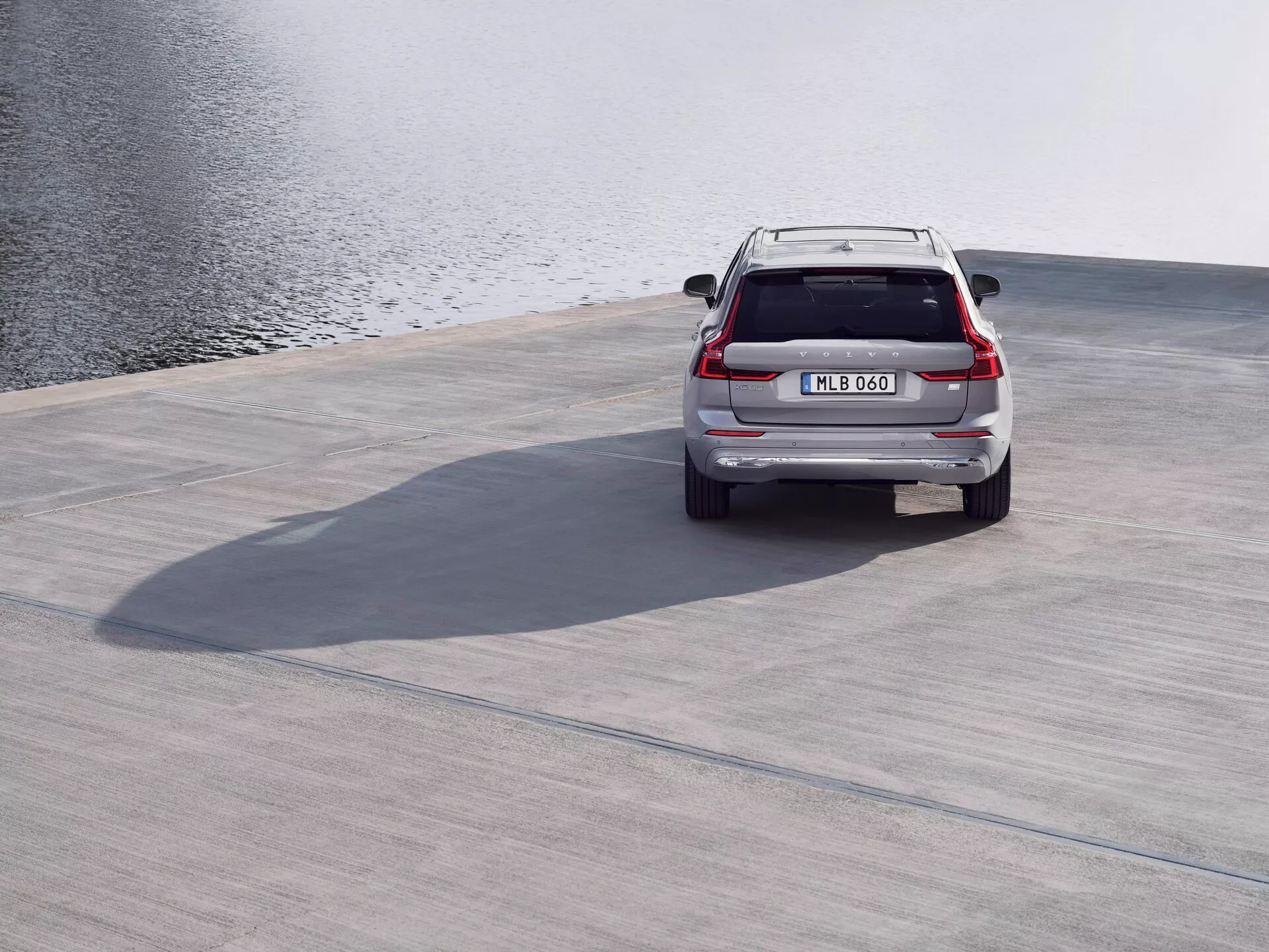 Pogled od zadaj na srebrni SUV Volvo XC60 2021 (druga generacija, prenova), parkiran na betonski površini v bližini vodnega telesa, s prikazanim strešnim spojlerjem, LED zadnjimi lučmi, prtljažnimi vrati, zadnjim odbijačem in izpušnimi cevmi iz visokega kota z metanimi sencami, v mirnih razmerah in lahkem vetru.