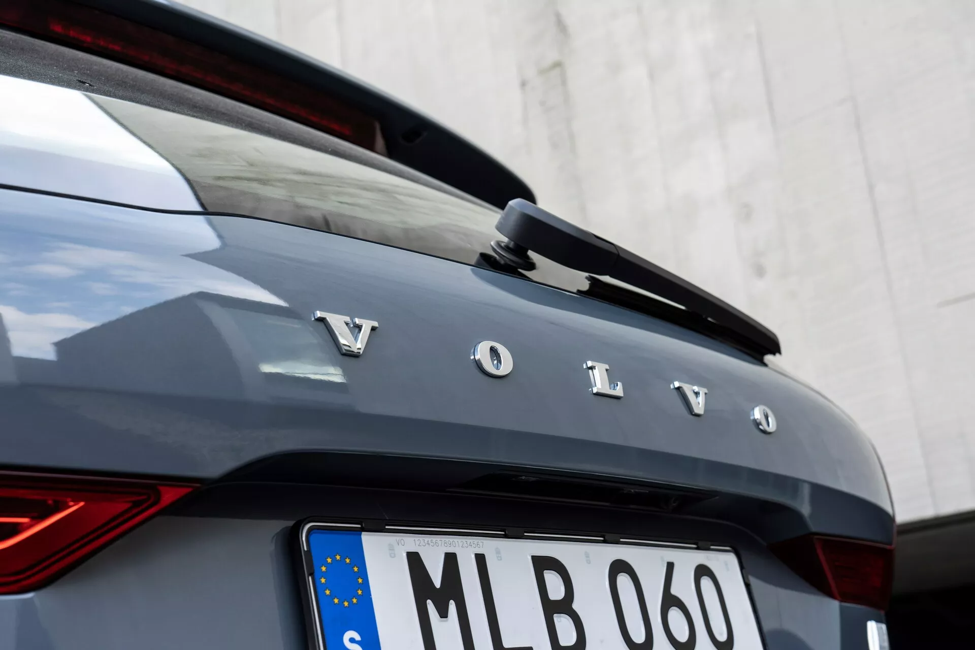 Podroben pogled na zadnji del avtomobila Volvo XC60 2021 (druga generacija facelift). Slika prikazuje debele, posamezne srebrne črke, ki sestavljajo 'Volvo' na zadnjem pokrovu, del zadnjega vetrobranskega stekla in integriran zadnji spojler s tretjo zavorno lučjo LED. Avto je temno sive ali grafitne barve, na levi strani je navpična LED zadnja luč z zasnovo ‘Thorjevo kladivo’. Pod imenom znamke je evropska registrska tablica z zastavo EU, ki pomeni registracijo v EU, obrobljena s kromirano obrobo. Kamera je rahlo nagnjena v desno, osredotoča se na črke in robove karoserije avtomobila, z nevtralnim belim ozadjem.