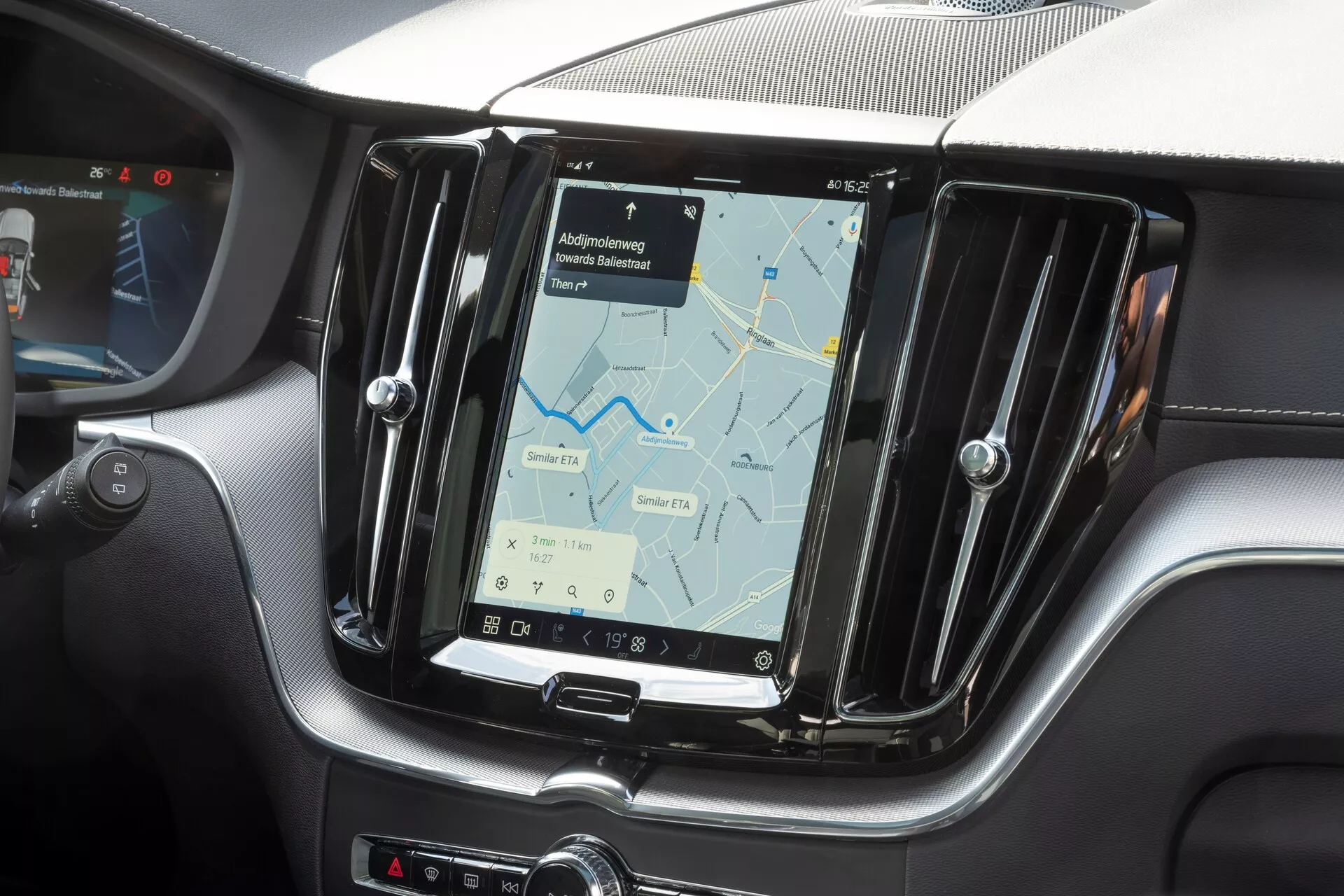 Podroben posnetek osrednje konzole v vozilu Volvo XC60 2021 (2. generacija, prenova), osredotočen na infotainment sistem. Fotografija, posneta z vidika sprednjega potnika, prikazuje velik navpični zaslon na dotik z navigacijskim zemljevidom. Zaslon je obrobljen s kovinskim okvirom in obdan s teksturo, podobno ogljikovim vlaknom, kar poudarja sodobne in športne oblikovalske elemente notranjosti. Fizični gumbi so vidni na strani zaslona in spodaj za nadzor klimatske naprave, kar kaže na kombinacijo funkcij na dotik in na dotik. Delni pogled na volan na levem robu potrjuje konfiguracijo vožnje na levi strani.