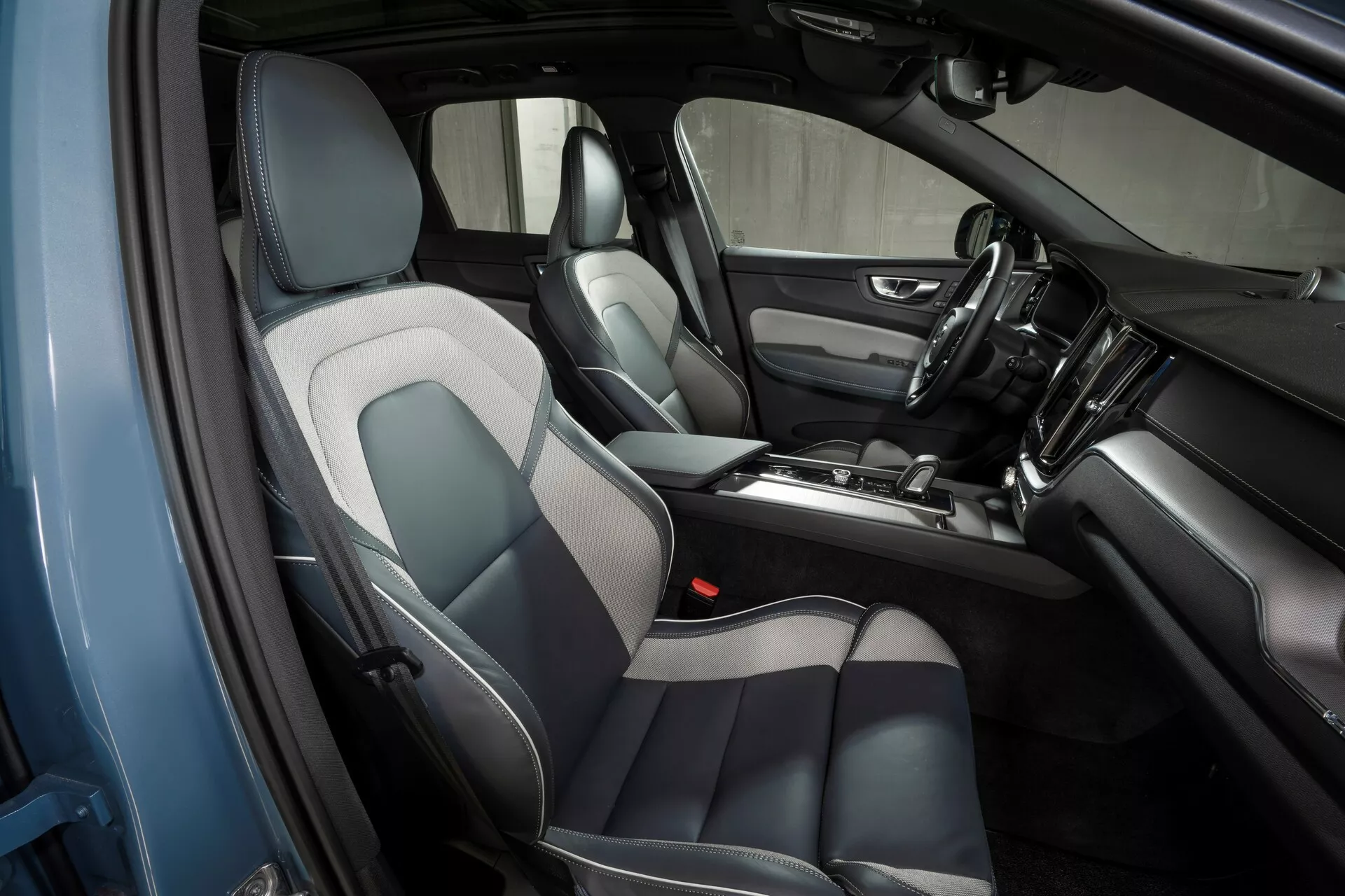 Notranjost vozila Volvo XC60 2021 (2. generacija-facelift), posneta iz dvignjenega, poševnega kota z voznikove strani, prikazuje zasnovo in značilnosti sprednje kabine, vključno z usnjenimi in tekstilnimi sedeži v sivi in črni barvi, sredinsko konzolo z prestavno ročico in različnimi kontrolami, armaturno ploščo z nameščenim zaslonom na dotik infotainment sistema, kontinuiteto v zasnovi vratnih plošč in vidnih mrežah zvočnikov.