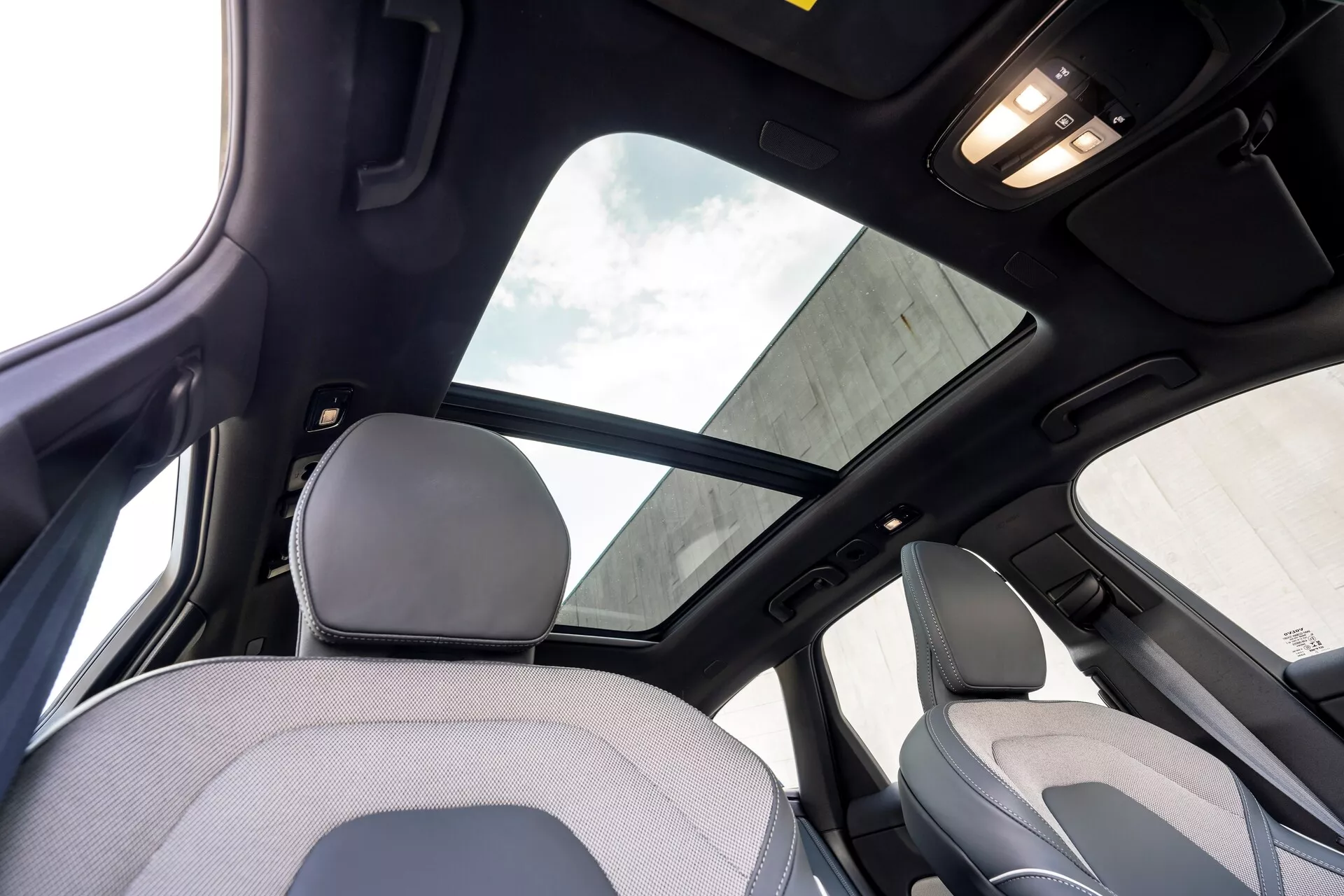 Notranjost vozila Volvo XC60 2021, facelift druge generacije, iz pogleda zadnjega sedeža, osredotočena na zgornji del kabine. Prepoznavne lastnosti vključujejo veliko panoramsko strešno okno s senčilom, črno strešno oblogo, ki označuje višjo stopnjo opreme ali luksuzni paket, ter osvetlitev kabine na stropu. Posnetek zajame del vzglavnika in zgornjega naslonjala sedeža za sovoznika, pa tudi B-stebriček in ročaj nad zadnjimi vrati.