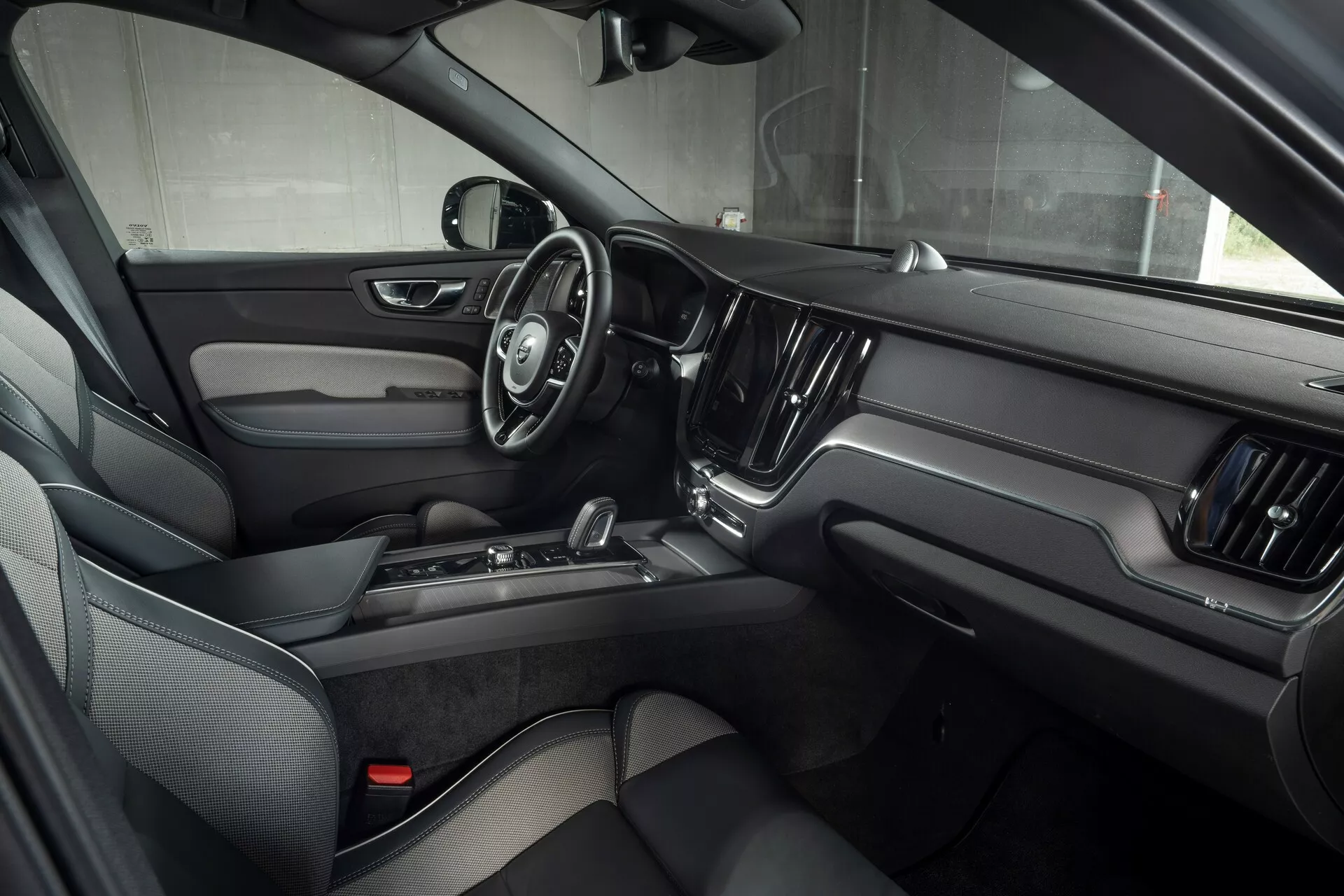 Notranji pogled na Volvo XC60 2021 (2. generacija facelift) s sedeža prednjega sopotnika, poudarjajoč voznikova vrata z elektronskimi kontrolnimi sistemi, trikraki volan z večnamenskimi gumbi, velik osrednji zaslon na dotik za informacijsko-razvedrilni sistem ter luksuzen dizajn s temnim usnjem in kovinskimi poudarki.
