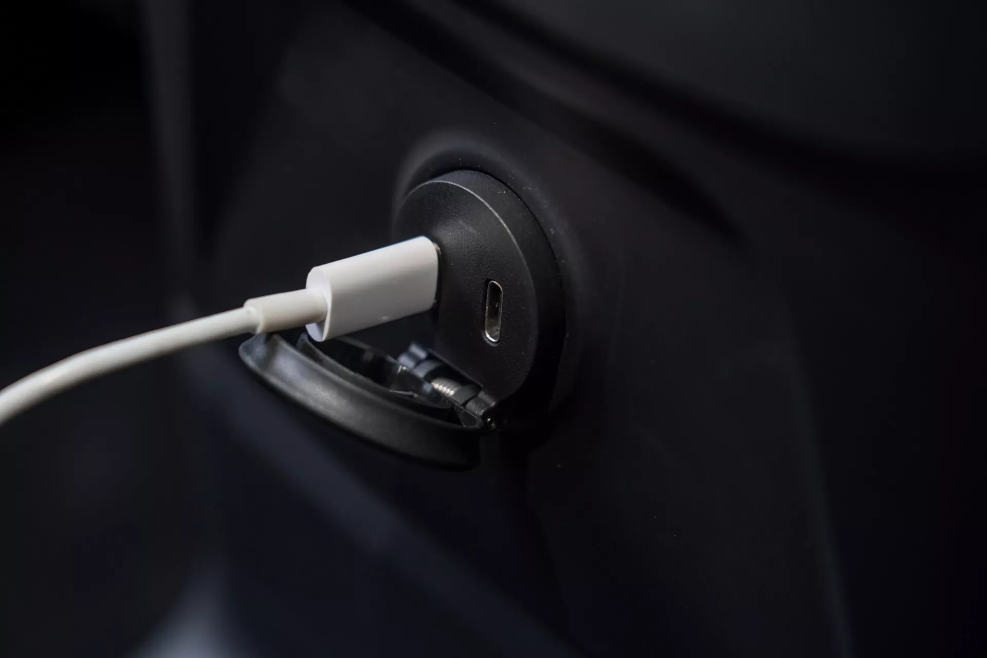 Od blizu prikazuje polnilni priključek električnega vozila Volvo XC60 2021 s priključenim kablom, poudarja eleganten in sodoben dizajn. Kadriranje se osredotoča na polnilni priključek, večji del temnega telesa vozila pa je večinoma neviden. Zamegljeno ozadje poudarja natančnost EV postopka polnjenja, kar simbolizira zeleno tehnologijo in trajnostni prevoz.