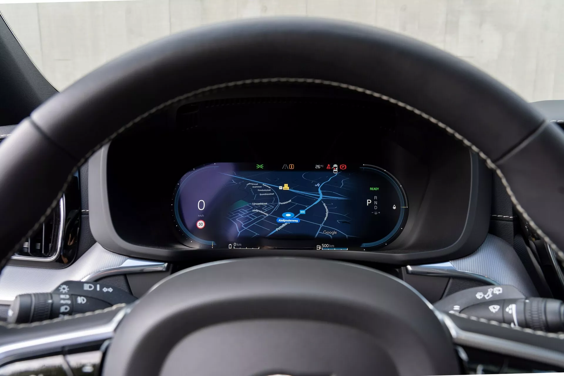 Pogled od blizu na armaturno ploščo Volvo XC60 2021 (2. generacija-facelift) iz perspektive voznikovega sedeža, osredotočena na digitalni instrumentni zaslon z usnjenim volanom z detajli šivov in večfunkcijskimi gumbi za medije, glasovne ukaze, tempomat in sisteme za pomoč vozniku. Digitalni zaslon prikazuje zemljevid z označeno navigacijsko potjo, merilnik hitrosti, tahometer in izbiro prestav v parkirnem načinu, obdan s premium notranjimi materiali.