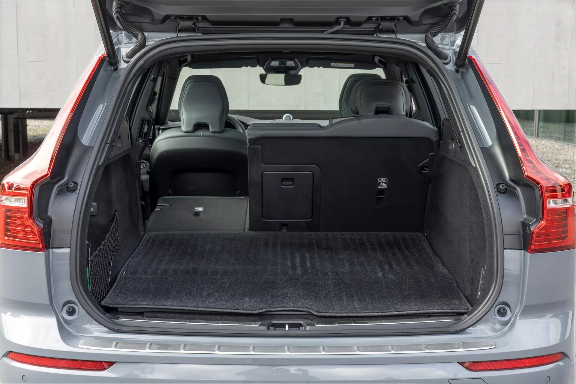Slika prikazuje zadnji tovorni prostor vozila Volvo XC60 2021 (2. generacija facelift), gledano iz ravnega kota z odprtim prtljažnikom. Srednji doseg zajame celotno širino tovornega prostora, ki ima ravno nakladalno dno, prekrito s črnim preprogi podobnim materialom. Na levi strani je majhen odlagalni predal z mrežico. Desni zadnji sedež je zložen, kar razkriva razdeljen vgrajen sedež, medtem ko levi sedež ostaja pokončen z vzglavnikom. Retraktibilna pokrov prtljažnika je nameščen nad zadnjimi sedeži, kar ponazarja vozilo vsestranskost skladiščenja.