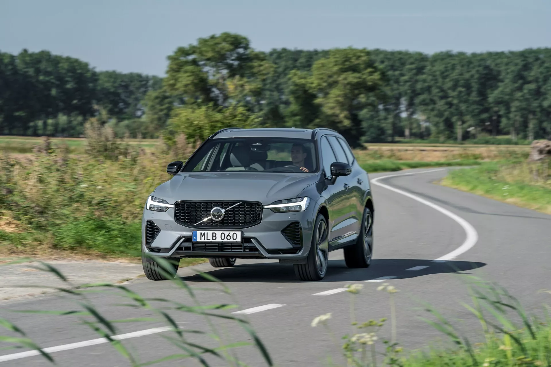 Slika z odseva Volvo XC60 2021 (osvežitev 2. generacije) v gibanju iz sprednjega trikotnega kota, poudarja oblikovne elemente, kot so vidna mreža s centralno postavljenim logotipom, kotni LED žarometi in elegantne linije pokrova. Avto, ki vozi po rahlo zaviti cesti, ima kovinsko barvo in večkraka alu platišča, obkroženo z oblikovanimi linijami in stranskimi ogledali z vgrajenimi smerniki. V ozadju so polja, drevesa in jasno modro nebo, ta predstavitev izpostavlja avtomobilski dizajn, cestna potovanja in resnične vozne scenarije.