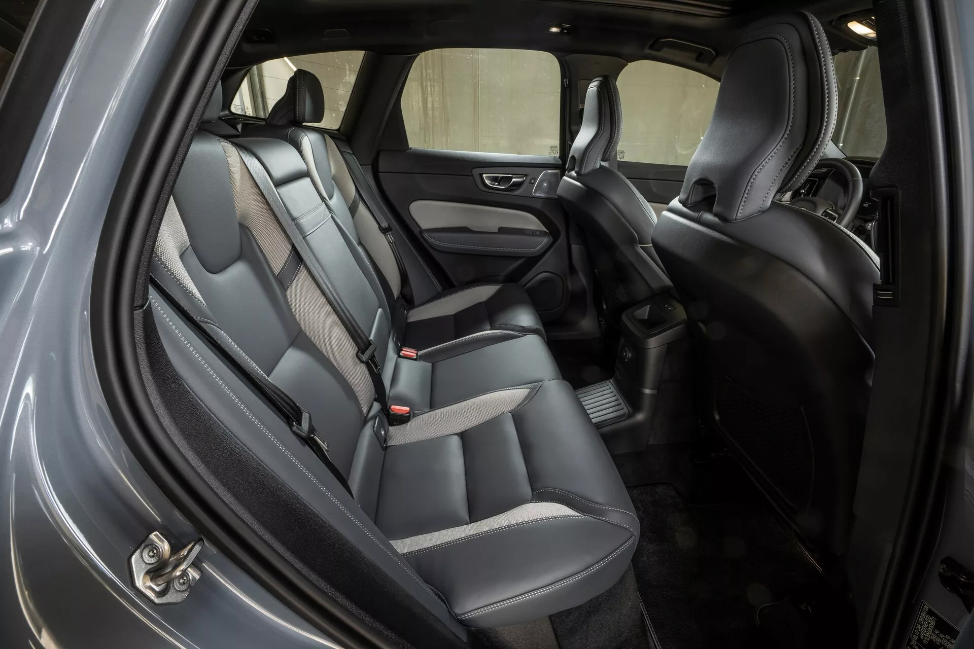 Posnetek notranjosti Volva XC60 2021, ki prikazuje zadnji sedežni prostor pod kotom s odprtimi zadnjimi vrati na pločniku. Avto ima usnjene ali imitacije usnja dvobarvne sedeže, naslonjala za glavo, zaponke varnostnih pasov za tri potnike in oblazinjene panele vrat z vgrajenim zvočnikom in nadzornimi gumbi za električna okna. Tla so prekrita s preprogo s podlogo, med sprednjimi sedeži je sredinska konzola, ki vključuje prostor za shranjevanje. Dobro osvetljena kabina nakazuje ambientalno in nadstropno osvetlitev ter ponuja dobro vidljivost skozi stranska in zadnja okna.