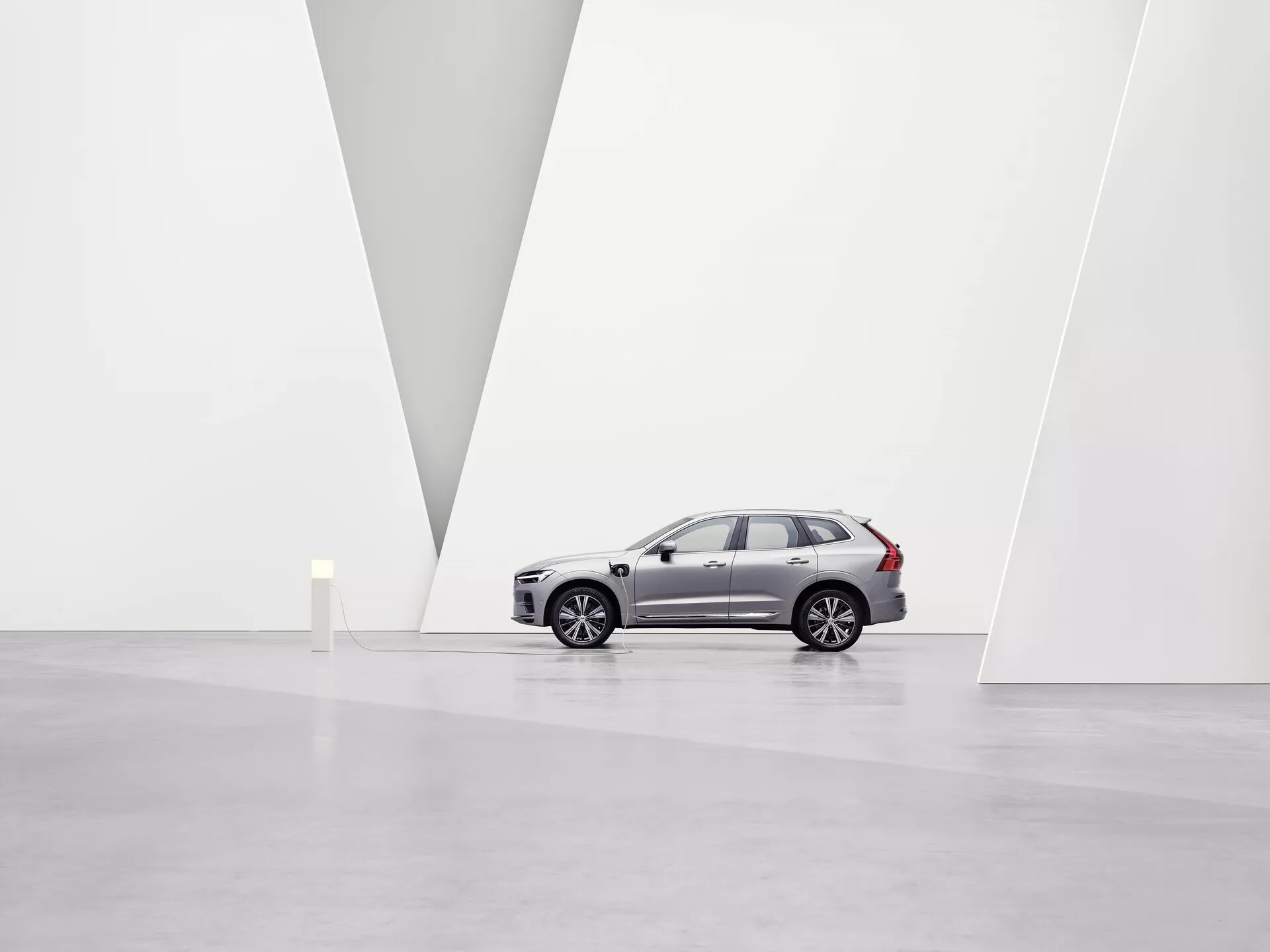 Slika prikazuje Volvo XC60 2021 druge generacije facelift SUV v prostorni minimalistični umetniški galeriji z belimi stenami in sijočimi tlemi. Vozilo, prikazano v profilu proti levi, poudarja aluminijasta kolesa, značilne linije na stranskih vratih, stranska ogledala, zatemnjena stekla in značilne strešne nosilce. Iz rahlega nizkega kota so vidne sprednja rešetka, LED žarometi, integrirani smerniki in parkirni senzorji na sprednjem odbijaču. Čisto okolje in V-oblikovana pregrada na steni poudarjata dizajn avtomobila in njegovo premium pozicijo na trgu pod mehko, razpršeno osvetlitvijo.