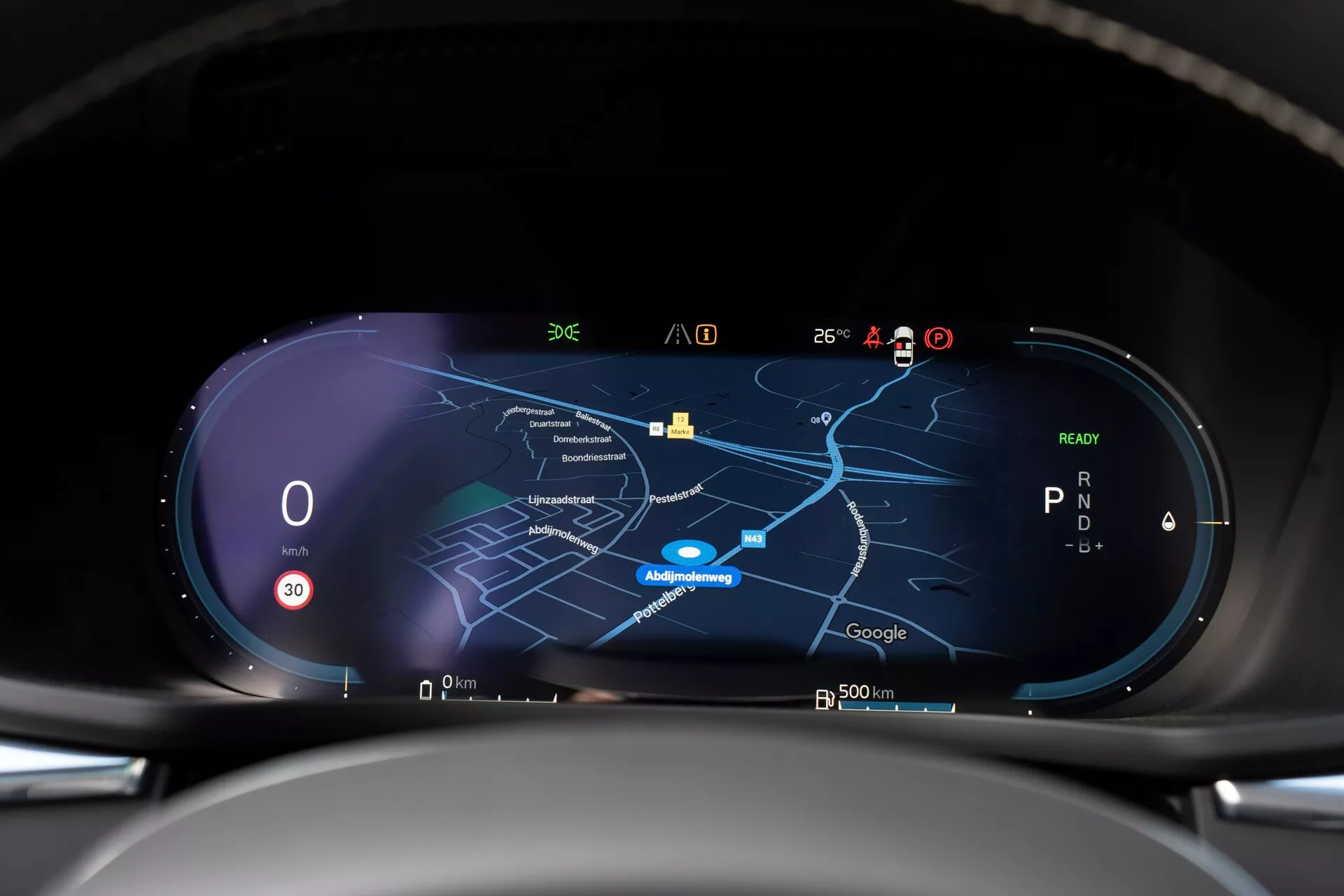 Notranji pogled Volvo XC60 2021, druge generacije z faceliftom, prikazuje visokoločljivostni digitalni instrumentni panel iz voznikove perspektive. Instrumentna plošča ima širokozaslonski prikaz s hitrostnim merilnikom, ki kaže ničlo, kazalnikom prestav, ki prikazuje 'P', in osrednjim navigacijskim zemljevidom v nočnem načinu. Različni ikoni za sprejem signala, zunanjo temperaturo, stanje baterije in funkcije pomoči vozniku so vidni vzdolž zgornjega roba, kar nakazuje na povezljivost v realnem času in napredne tehnološke zmogljivosti.