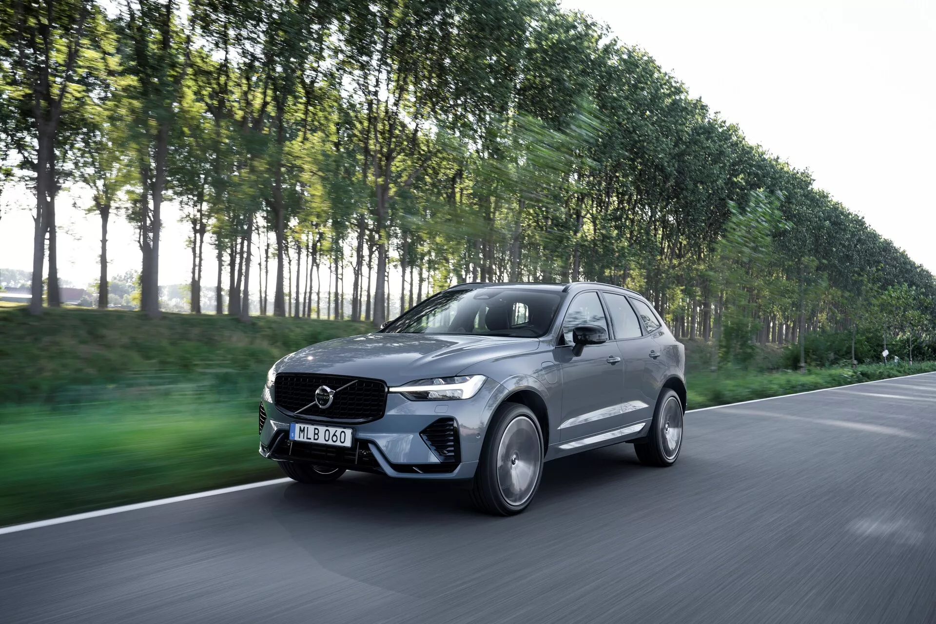 Na sliki je prikazan premikajoči se Volvo XC60 2021 (prenova 2. generacije) zajet iz kota s treh četrtin spredaj, ki kaže voznikovo stran avtomobila. Ta sodoben luksuzni SUV ima eleganten dizajn in viden emblem na sprednji rešetki. Posnetek s rahlim spuščenim kotom vključuje pokrov motorja, odbijač, rešetko, žaromete in kolesa. Temno kovinsko vozilo, verjetno sivo ali črno, s sijajnim zaključkom, ima platišča iz zlitine, LED žaromete in aerodinamično karoserijo. Zamegljeno ozadje ulične pokrajine z drevesi nakazuje gibanje in poudarja zmogljivosti avtomobila.