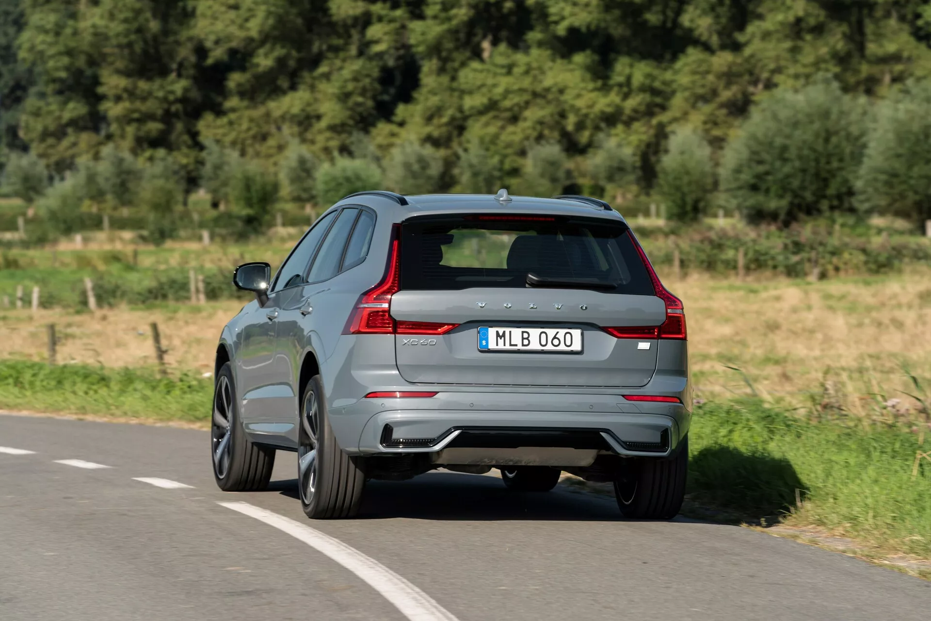 Zadnji pogled na Volvo XC60 2021 iz treh četrtin, ki se premika po asfaltirani cesti. Posnetek iz zvišanega kota prikazuje ta sodobni SUV s sijočo sivo barvo, značilnimi LED zadnjimi lučmi, dvojnimi izpušnimi cevmi in zadnjim difuzorjem. Znak proizvajalca in oznaka modela sta vidna na oblikovanih zadnjih vratih, ki jih dopolnjujejo zatemnjena okna in litinska platišča z večkrakovim dizajnom, kar nakazuje na bolj športno različico. Slika ponuja jasen pogled na aerodinamični dizajn in vrhunske lastnosti, značilne za sodobna vozila.