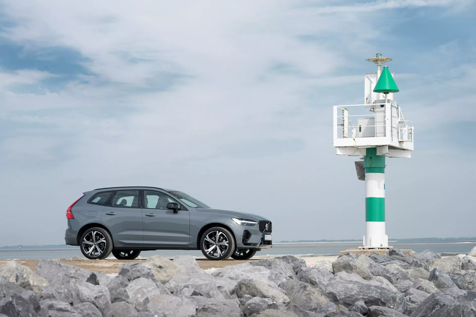 Profilski pogled na siv SUV Volvo XC60 2021 (druga generacija, facelift) na kamnitem terenu z morskim svetilnikom v ozadju. Eleganten dizajn vključuje kromirano obrobo okenskih stekel, večkrake aluminijaste platišča in integriran smernik v stranskem ogledalu, ujete proti delno oblačnemu obalnemu nebu.