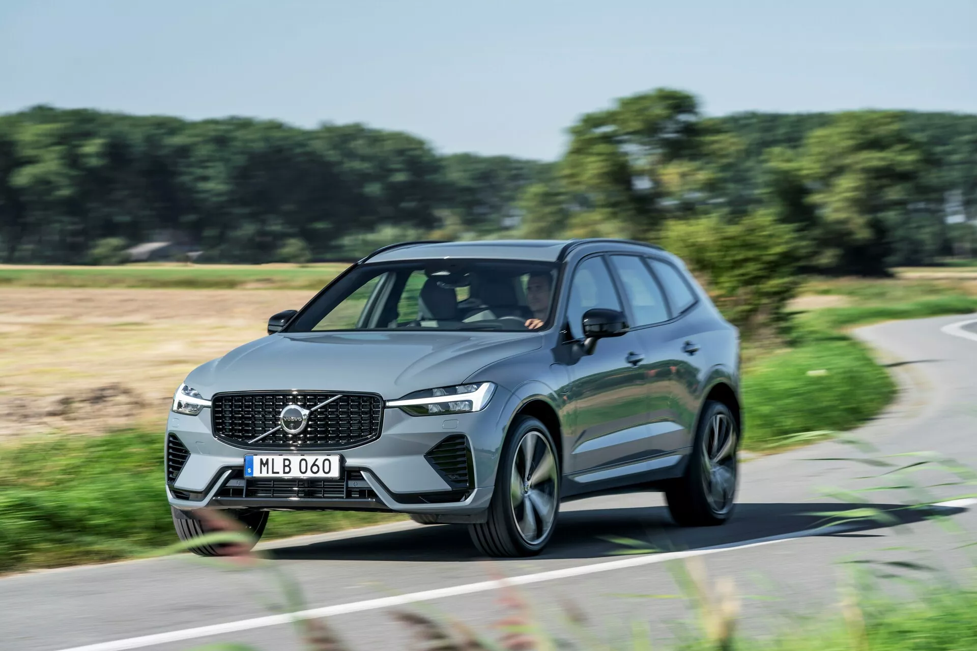 Dinamičen dnevni posnetek premikajočega se vozila Volvo XC60 2021 (druga generacija-facelift) SUV, zajet z tridčetrtglednega prednjega kota. Slika prikazuje sprednjo mrežo z izrazitim emblema Volvo, kotne LED žaromete, aerodinamične črte in sodobna večkraka aluminijasta platišča. Dnevne luči so prižgane, kar nakazuje, da je avto v gibanju. Fotografija poudarja eleganten dizajn in prisotnost na cesti tega sodobnega družinskega ali luksuznega vozila.