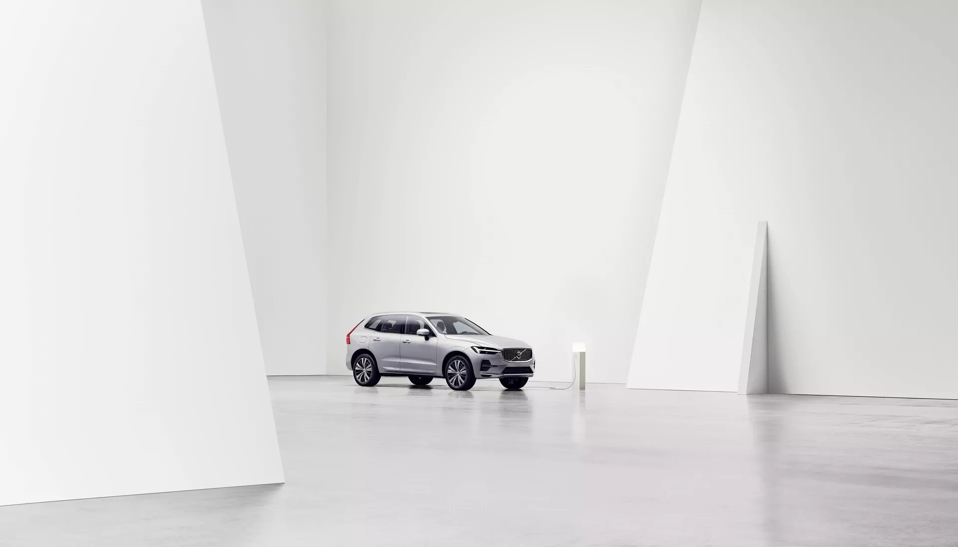 Slika Volva XC60 2021 (2. generacija, facelift), postavljenega centralno v minimalističnem, prostornem, dobro osvetljenem studijskem okolju, posneta iz sprednje tročetrtinske perspektive. Svetlo srebrna barva avtomobila kontrastira s temnejšimi kolesi in delom sprednje maske, ki prikazuje sodobne oblikovalske elemente SUV, kot so ukrivljene in kotne linije. Pogled poudarja sprednjo masko, žaromete, pokrov motorja, vetrobransko steklo in stran voznika, vključno s sprednjimi in zadnjimi vrati ter stranskimi okni. Čisto belo ozadje poudarja avto, usmerja pozornost na njegov dizajn in lastnosti, primerno za promocijske ali umetniške namene.