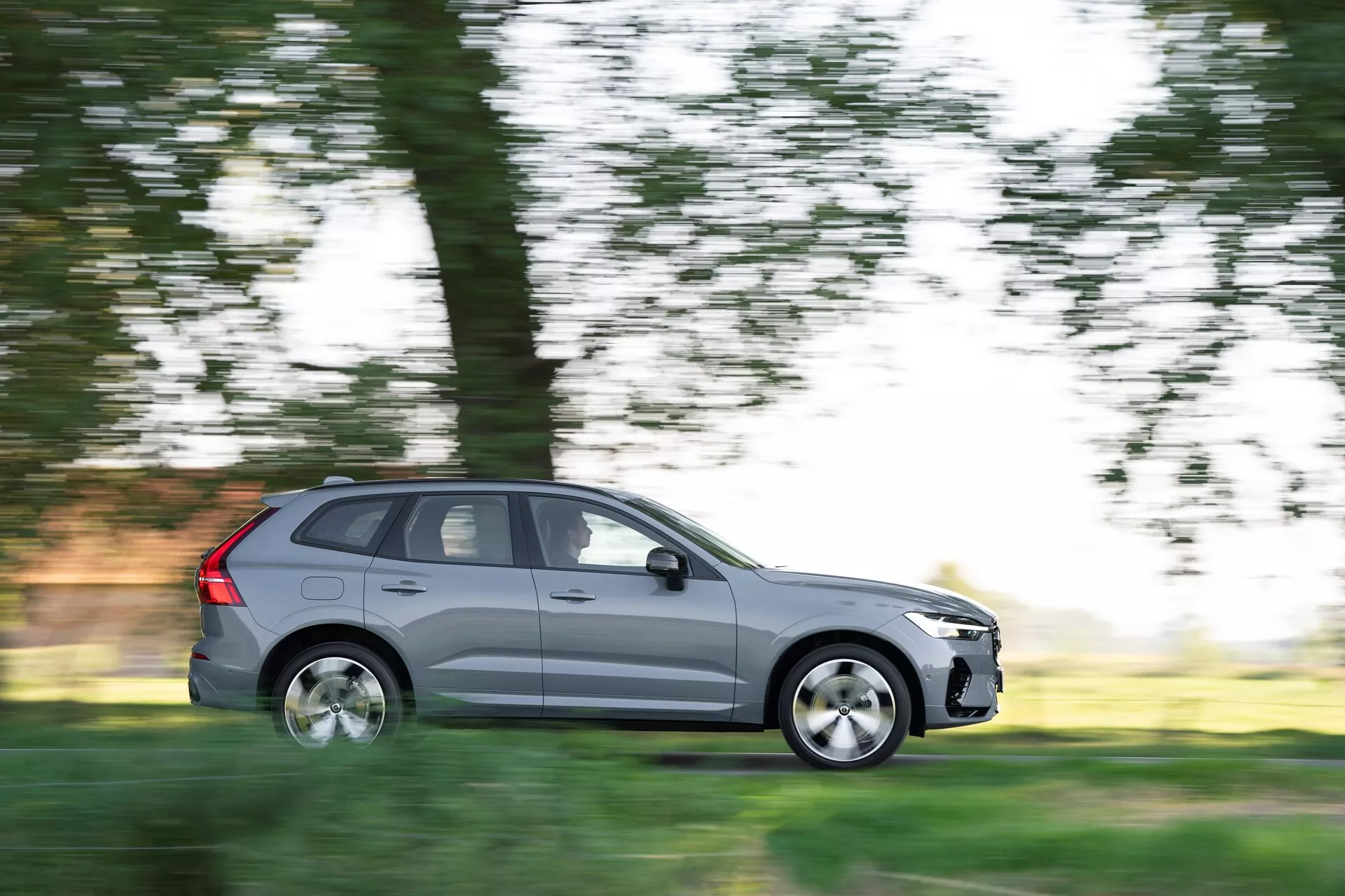 Slika prikazuje SUV Volvo XC60 2021 (2. generacija, facelift) v gibanju, zajeto s tehniko panoramskega slikanja, ki zamegli ozadje, medtem ko avtomobil ostane ostro v fokusu. Prikazan s strani voznika v celotnem bočnem profilu, kovinsko siv vozil ima pet vrat, litkocestna kolesa in standardne pnevmatike. Prizor nakazuje vožnjo podnevi v naravnem okolju, z meglenimi drevesi in travo v ozadju, kar poudarja gibanje avtomobila.