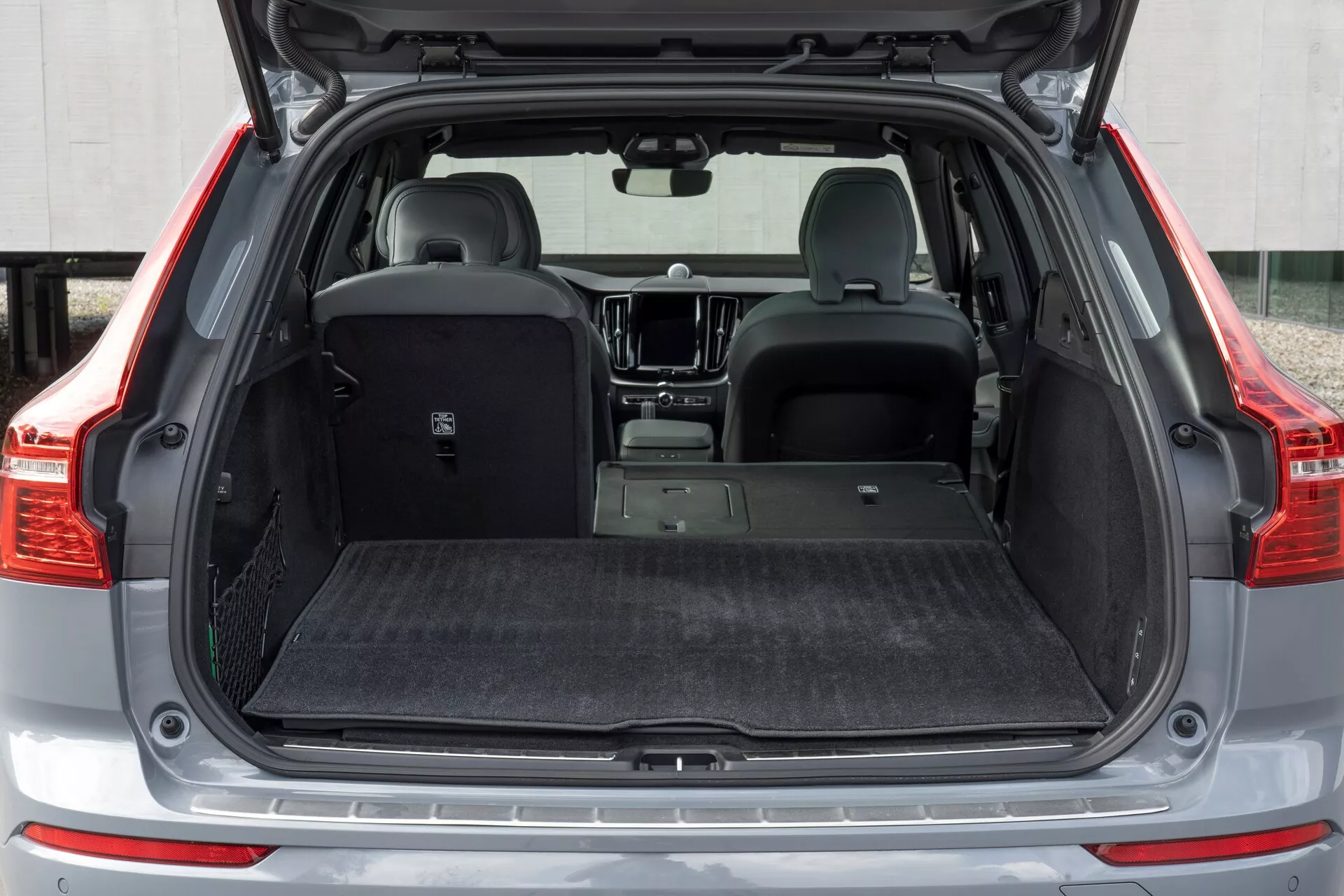 Pogled od zadaj od blizu na drugo generacijo prenovljenega modela Volvo XC60 2021, ki prikazuje odprt prtljažnik iz nizkega kota. Slika poudarja prostoren tovorni prostor z minimalnim vdorom koles, izvlečno prtljažno pokrivalo in oblazinjeno notranjost, usmerjeno na zmanjševanje hrupa in zaščito. Delno zloženi zadnji sedeži prikazujejo vsestranskost, medtem ko zaščitna plošča na nakladalnem pragu, vidni parkirni senzorji na zadnjem odbijaču, LED osvetlitev prtljažnika in sidrišča poudarjajo funkcionalnost in pozornost do podrobnosti.