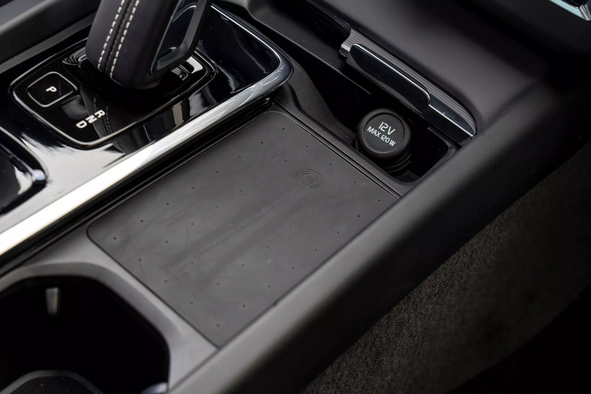 Zbližan, poševni pogled na območje sredinske konzole v vozilu Volvo XC60 2021 (druga generacija facelift) s potniške strani, z avtomatskim prestavnim ročajem v položaju 'park', gumiranim prostorom za shranjevanje, stikalom elektronske parkirne zavore, držali za kozarce in kombinacijo materialov iz mehke plastike in kovinskega zaključka v temno sivih in črnih tonih.