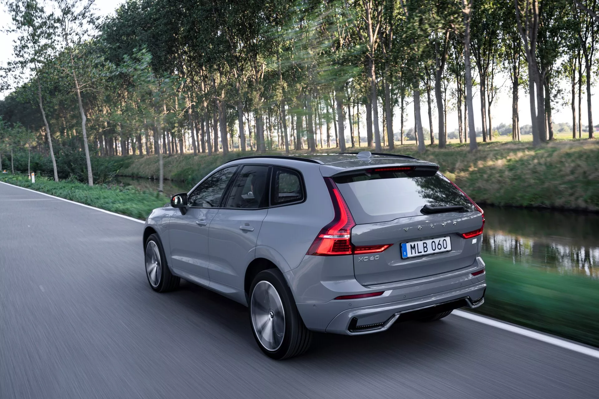 Dinamični zadnji trikotni pogled na sodoben SUV Volvo XC60 2021 (2. generacija-facelift) na asfaltirani cesti, poudarjajoč eleganten zadnji dizajn, vključno z zadnjimi lučmi, zadnjim oknom, prtljažnikom in linijo strehe. Slika, posneta iz nekoliko znižane perspektive, prikazuje stilizirana kolesa in bočne karakterske črte. SUV je prikazan v gibanju s zamegljeno naravno ozadje dreves in trave, kar kaže na parkovno nastavitev. Srebrno sivo luksuzno vozilo ima dvojni izpuh, aerodinamično zasnovo in sijočo obdelavo, z litimi platišči in verjetno napredno tehnologijo, kot so zadnje LED luči.