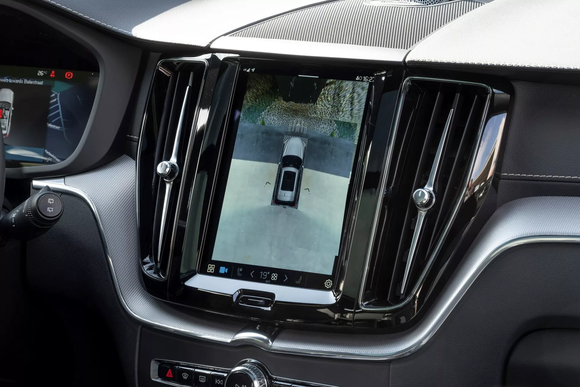 Notranji pogled na Volvo XC60 2021 (2. generacija s prenovo) z velikim, navpično usmerjenim infozabavnim zaslonom na dotik na sredinski konzoli, ki prikazuje sliko zadnje kamere z ptičje perspektive. Luksuzna površina s kromiranimi detajli in elegantnimi oblikovnimi elementi, dodatni kontrolniki pod zaslonom in delni pogled na volan, ki kažejo na vrhunsko in tehnološko napredno notranjost.