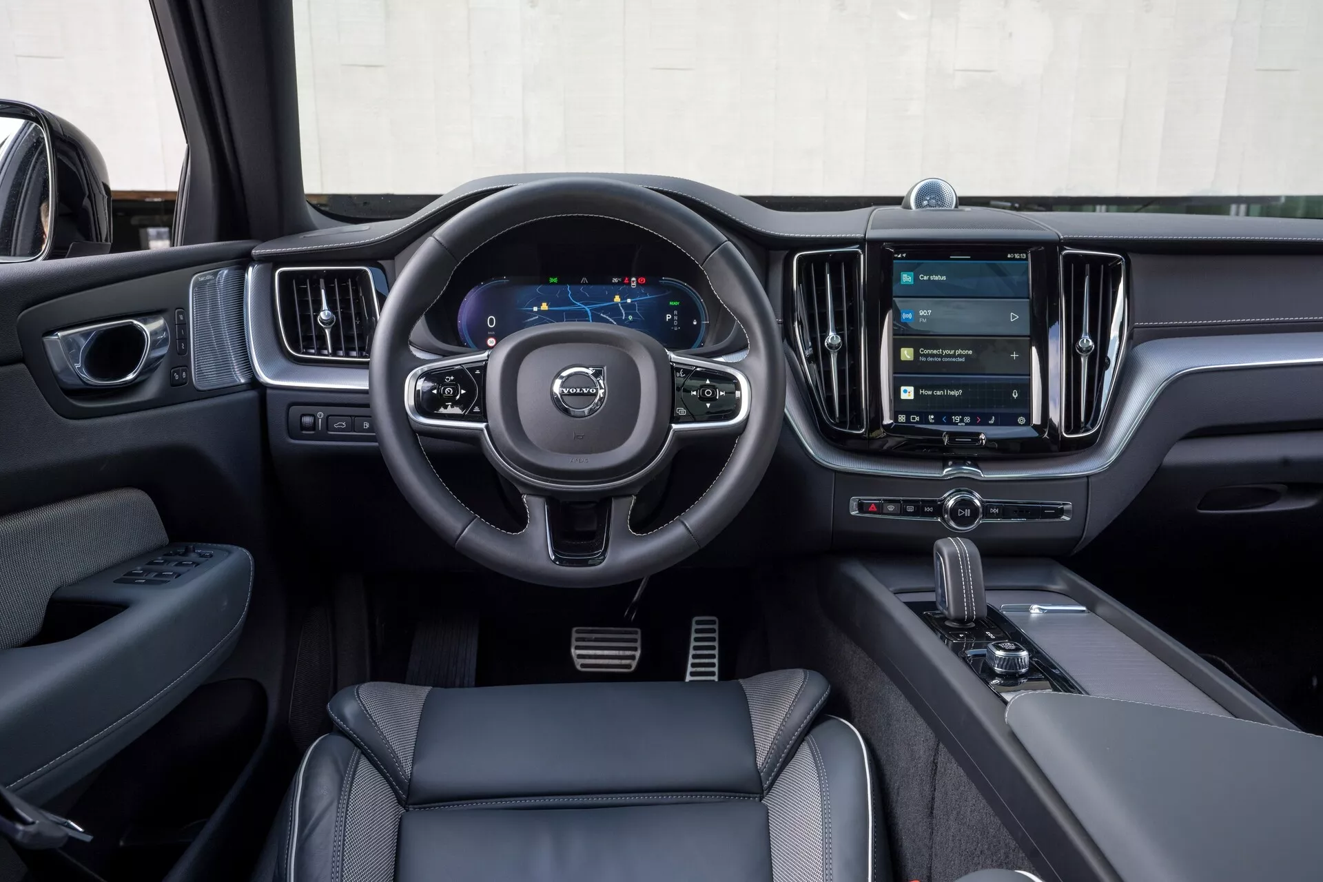 Notranjost vozila Volvo XC60 2021 (facelift druge generacije) iz perspektive voznika. Slika poudarja trikraki volan z gumbi za upravljanje zvoka, komunikacije in tempomata, digitalno armaturno ploščo, ki prikazuje merilnik hitrosti in navigacijo, ter velik vertikalno usmerjen zaslon na dotik infotainment sistema. Sodobni oblikovni elementi vključujejo usnjene ali usnju podobne materiale, kromirane dodatke ter prestavno ročico samodejnega menjalnika z izbirnikom načina vožnje. Na voznikovih vratih so vidni nadzorni paneli za nastavljanje oken in ogledal, kar poudarja tehnološke in luksuzne lastnosti.