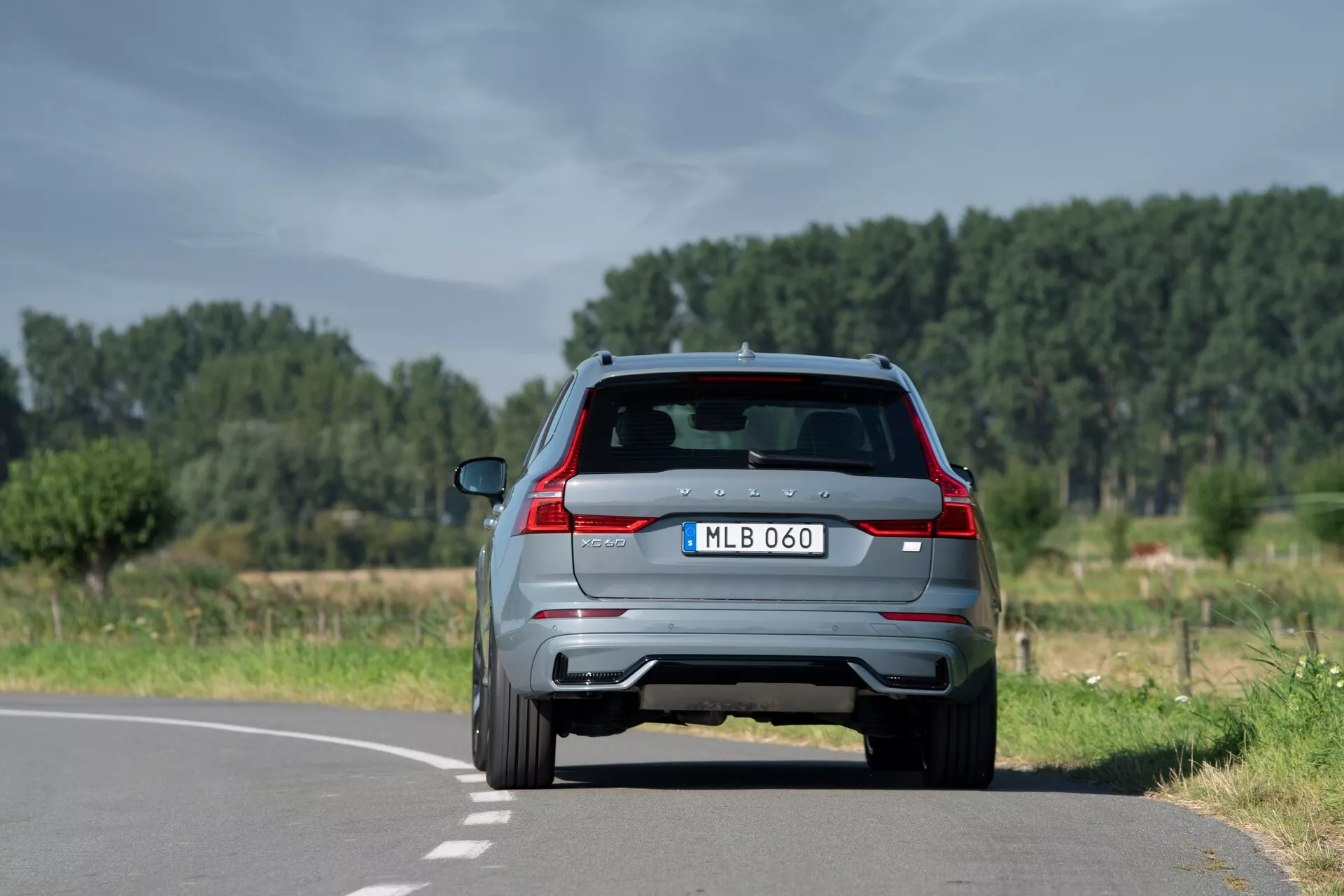 Zadnji pogled na sodoben SUV Volvo XC60 2021 (2. generacija-facelift) na dvopasovni cesti v naravnem, podeželskem okolju, ki prikazuje značilne zadnje luči, dvojni izpušni cevi, zadnji spojler z vgrajenimi zavorami, zadnjo kamero, zadnji brisalec in parkirne senzorje. Rahlo je postavljen na desno stran sredinske črte, z jasnim nebom in drevesi v ozadju.