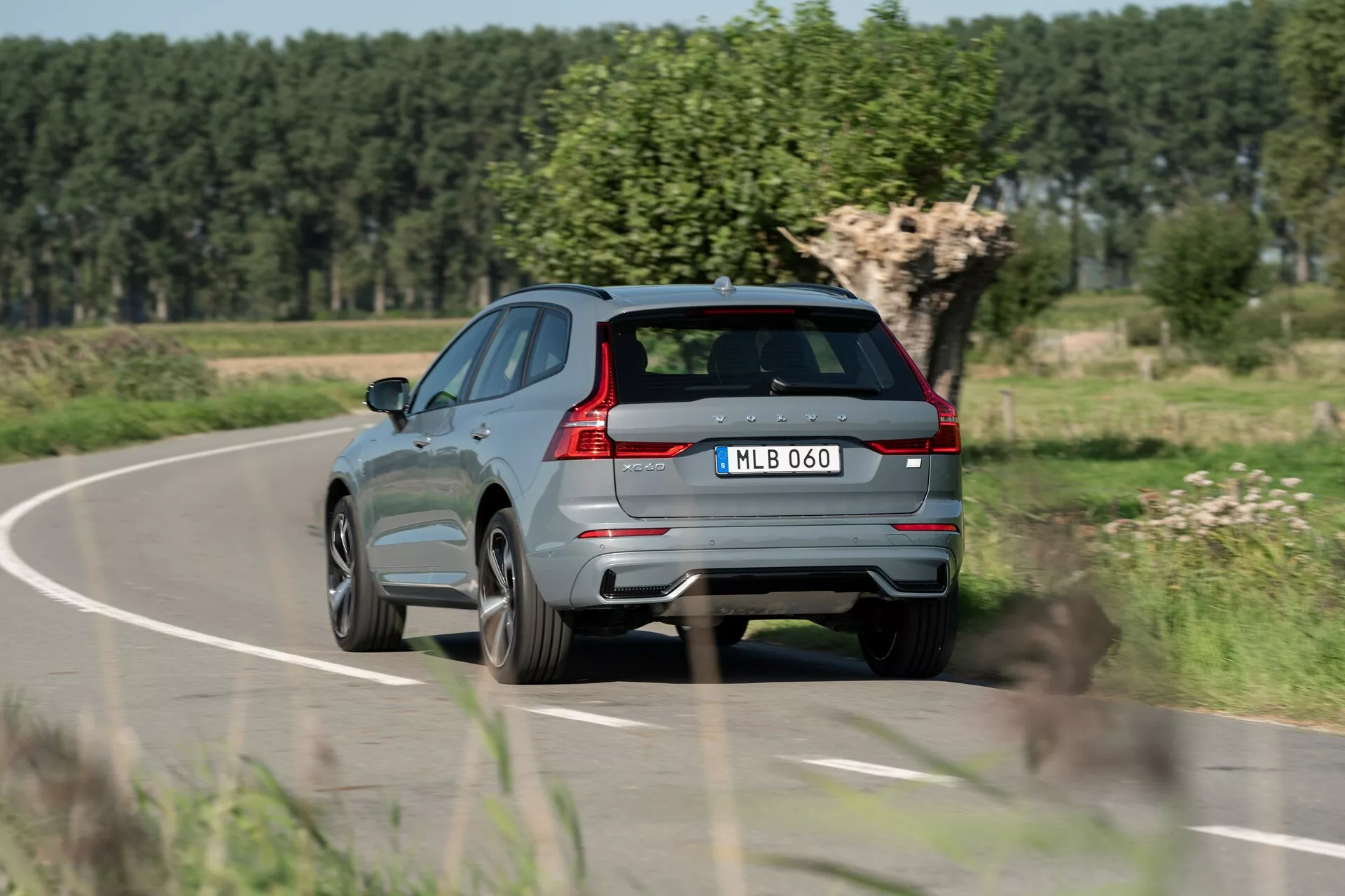 Zadnji tričetrtinski pogled na moderno kovinsko sivo SUV Volvo XC60 2021 v gibanju na podeželski cesti, prikazuje značilne zadnje luči, dvojni izpušni sistem, zadnji brisalec, izrazit zadnji spojler, integrirane senzorje na zadnjem odbijaču, tablica nameščeno centralno, aluminijasta platišča, ostro značilno črto vzdolž stranice, reflektivne obrobe oken in strešni sistem. Ozadje z zamegljenim listjem in jasnim nebom kaže na gibanje in podeželsko okolje.