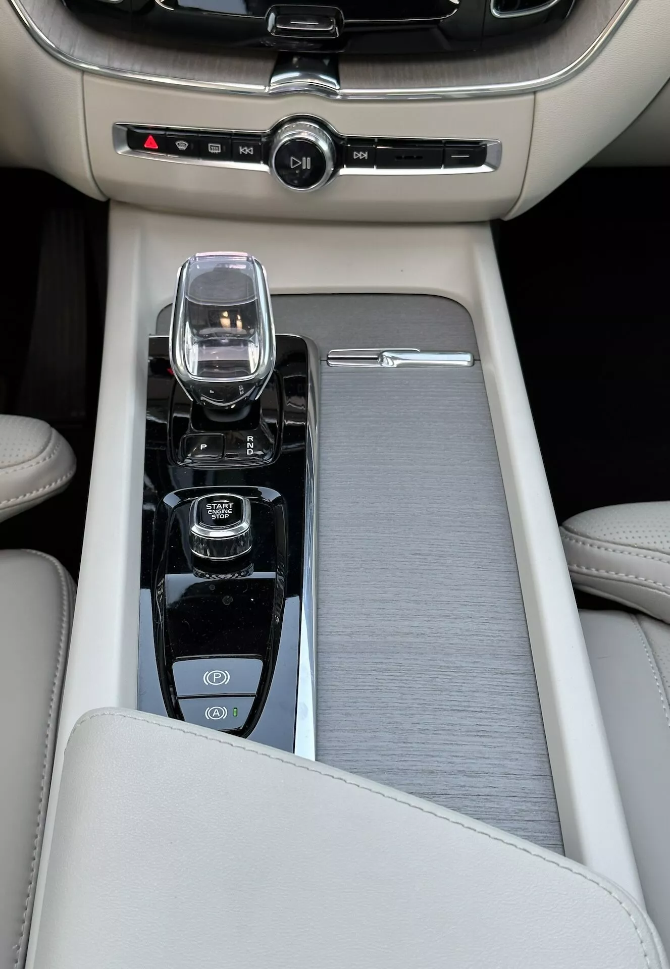 Podroben pogled na sredinsko konzolo v Volvo XC60 Recharge plug-in hibridu letnik 2021, z bež usnjenim naslonom za roke, drsnim kovinskim ali plastičnim okrasnim pokrovom, ki sega vzdolž celotne zgornje površine sredinske konzole, modernim krmilnikom za informacijsko-zabaviščni sistem, prestavno ročico iz kristalnega stekla, ki kaže na avtomatski menjalnik z elektronsko tehnologijo prestavljanja po žicah, in upravljalniki za start/stop motorja, izklop trakcijske kontrole in morda izbirnik načina za različne vozne dinamike, skupaj z nadzorom dvojne podnebne cone.
