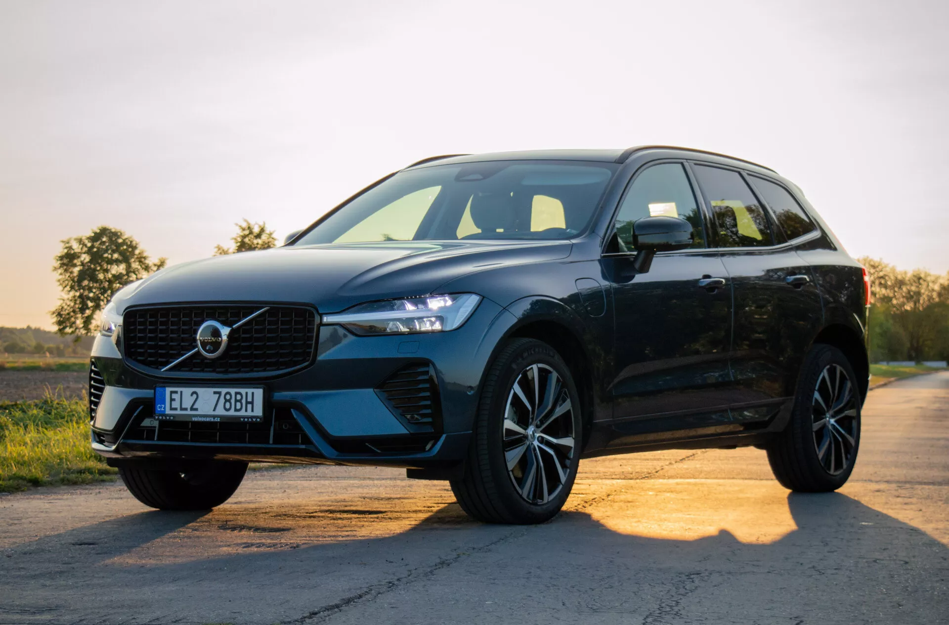Črni, sodobni SUV Volvo XC60 Recharge Plug-in Hybrid 2021 parkiran na asfaltni cesti ob sončnem vzhodu ali zahodu, prikazuje njegov dizajn s poudarkom na rešetki, LED žarometih, zlitinskih kolesih in stranskem profilu proti gradientnemu ozadju neba