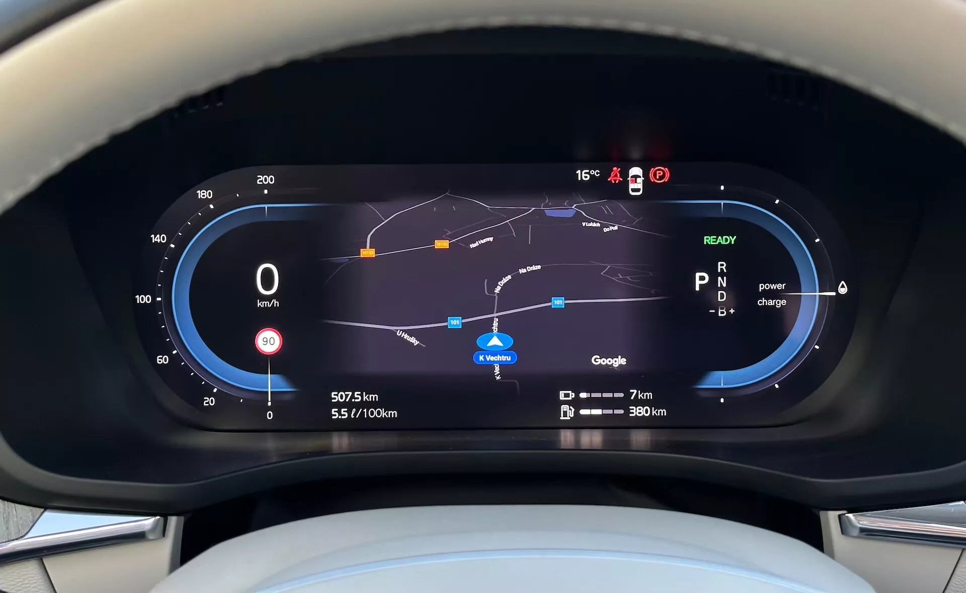Pogled na digitalni instrumentni panel priključnega hibrida Volvo XC60 Recharge 2021 iz perspektive voznika, ki vključuje merilnik hitrosti, merilnik učinkovitosti goriva, porabo moči, navigacijski zemljevid in različne indikatorje vožnje