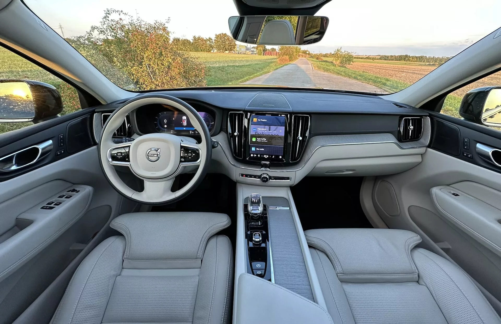Pogled na notranjost Volva XC60 Recharge Plug-In Hybrid 2021, vzeto iz zadnjega sedežnega prostora, prikazuje celotno armaturno ploščo, večfunkcijski volan z logotipom, digitalno instrumentno ploščo, centralni zaslon na dotik sistema za informacije in zabavo ter luksuzne sedeže iz svetlega usnja.