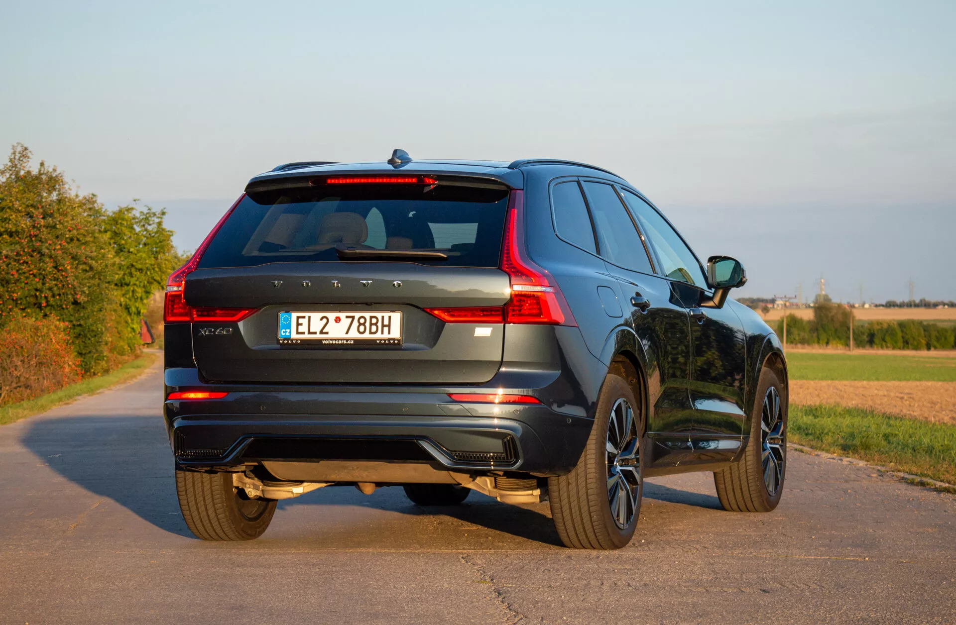 Stacionarni SUV Volvo XC60 Recharge plug-in hibrid iz leta 2021 na tlakovani cesti, ki poudarja svoj pogled s treh četrtin zadaj, osvetljuje LED zadnje luči, zlitinska kolesa in dinamičen dizajn v podeželskem okolju pod jasnim nebom
