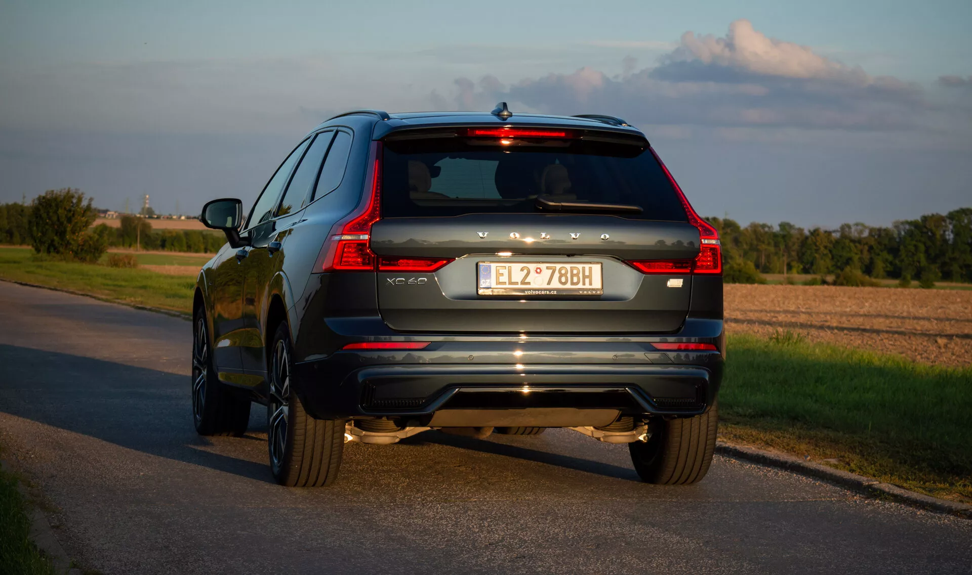 SUV Volvo XC60 Recharge priključni hibrid 2021 na asfaltni cesti, posnet z zadnjega kota tri četrtine, ki poudarja LED zadnje luči vozila, izpušni sistem in kromirane poudarke v poznem popoldnevu