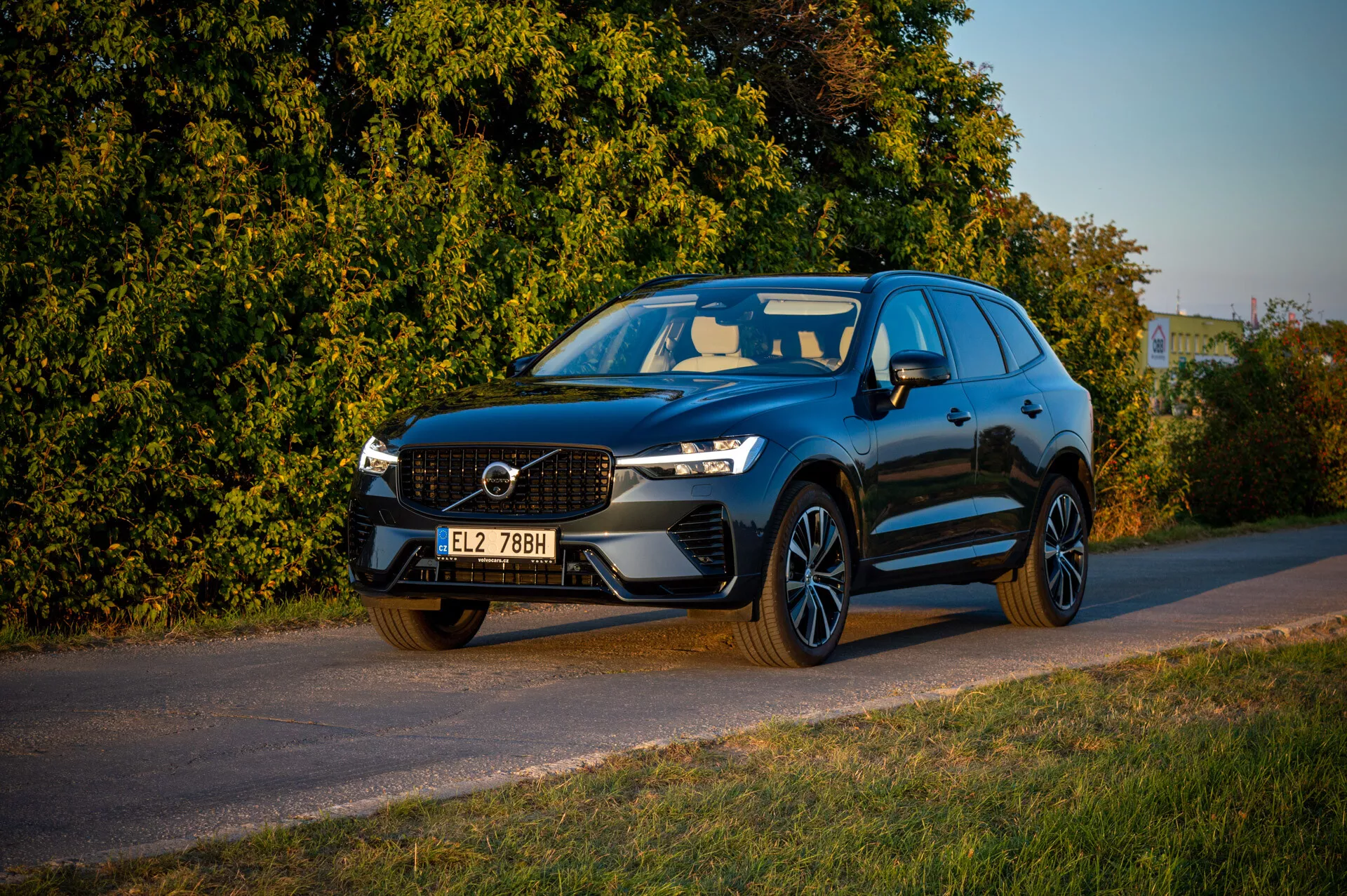 Volvo XC60 Recharge Plug-In Hybrid iz leta 2021 parkiran ob cesti v poznem popoldnevu s toplo osvetlitvijo, prikazuje sprednjo rešetko, LED žaromete in športni profil na ozadju vegetacije