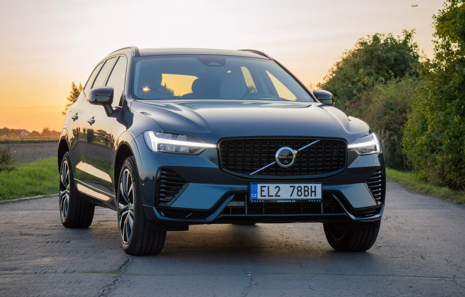Luksuzni SUV Volvo XC60 Recharge Plug-In Hybrid 2021 na asfaltirani cesti z naravnim ozadjem med zlato uro, prikazujejoč sprednjo masko, LED žaromete, oblikovan pokrov motorja, večkraka lita kolesa in svetlečo površino