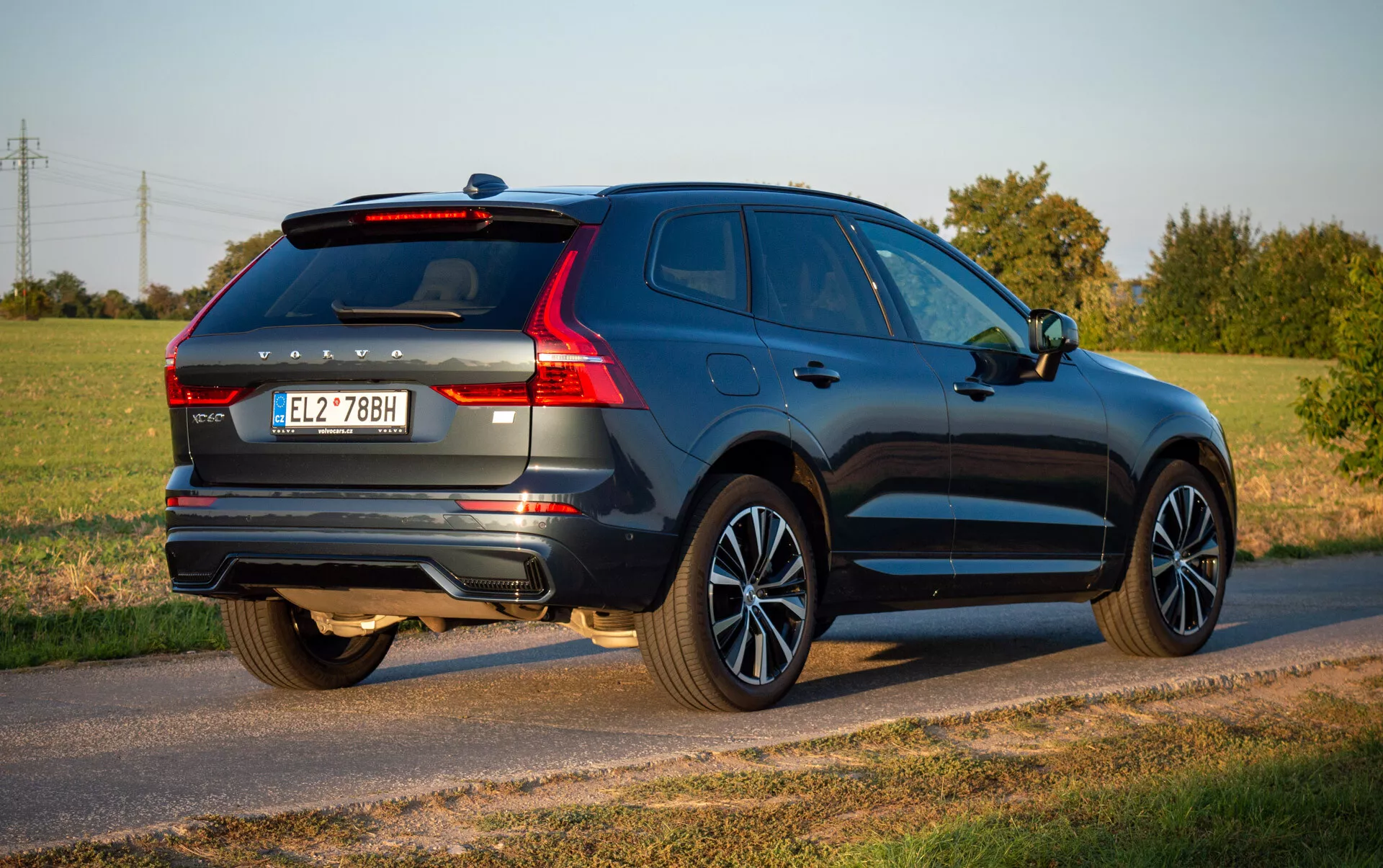 Volvo XC60 Recharge Plug-In Hybrid letnik 2021 v globoko modri barvi, ki prikazuje aerodinamično obliko in sodobne značilnosti kot so LED zadnje luči, litna platišča in dvojni izpušni sistem, parkirano na podeželski cesti pod jasnim nebom