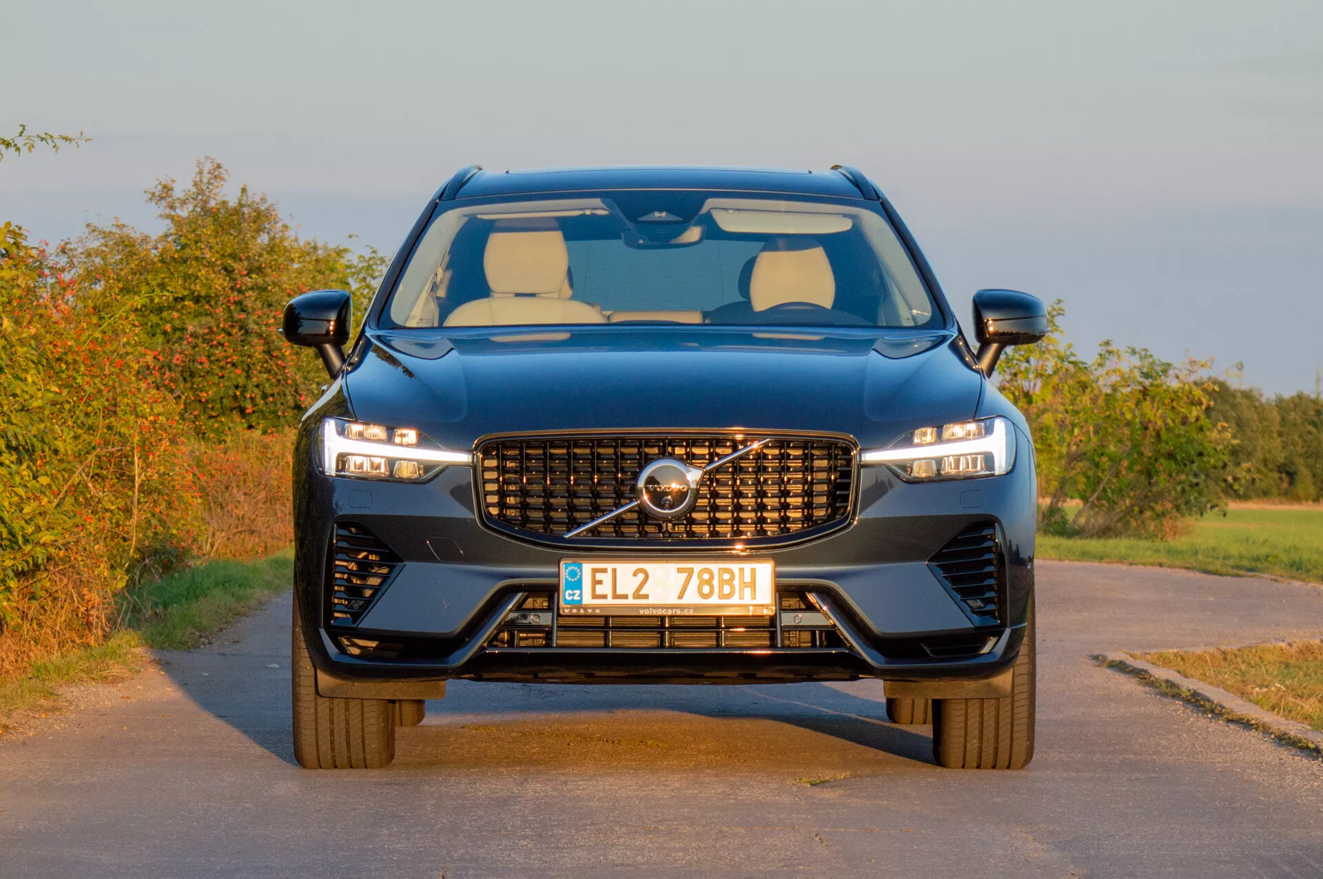 Volvo XC60 Recharge Plug-In Hybrid 2021, ki prikazuje svoj sodobni dizajn z značilno rešetko, elegantnimi LED žarometi in športnim sprednjim odbijačem na podeželskem ozadju v topli svetlobi poznega popoldneva