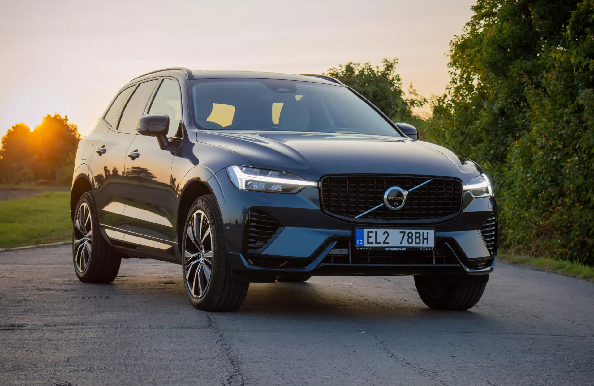 Volvo XC60 Recharge Plug-In Hybrid 2021 v moderni zasnovi SUV, rahlo nagnjeno za izpostavitev sprednjega četrtletja, kar prikazuje značilno rešetko, elegantne LED žaromete in aerodinamični sprednji odbijač z integriranimi zračnimi režami, dopolnjeno z sijočim zunanjim zaključkom in premijskimi večkrakimi zlitimi kolesi pod naravno dnevno svetlobo.